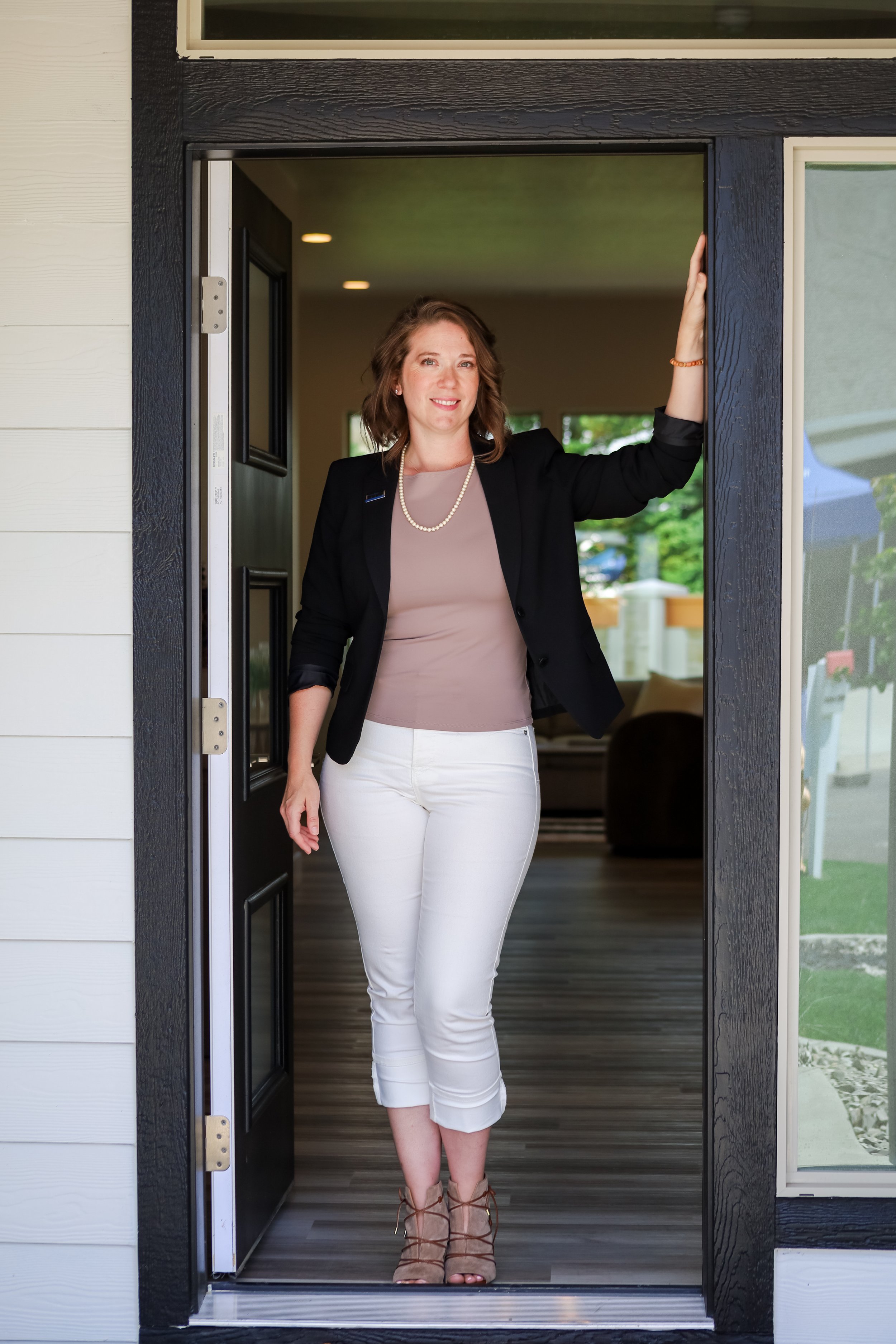 A woman in a black blazer, beige top, white capri pants, and beige heels standing in a home's front door, smiling.