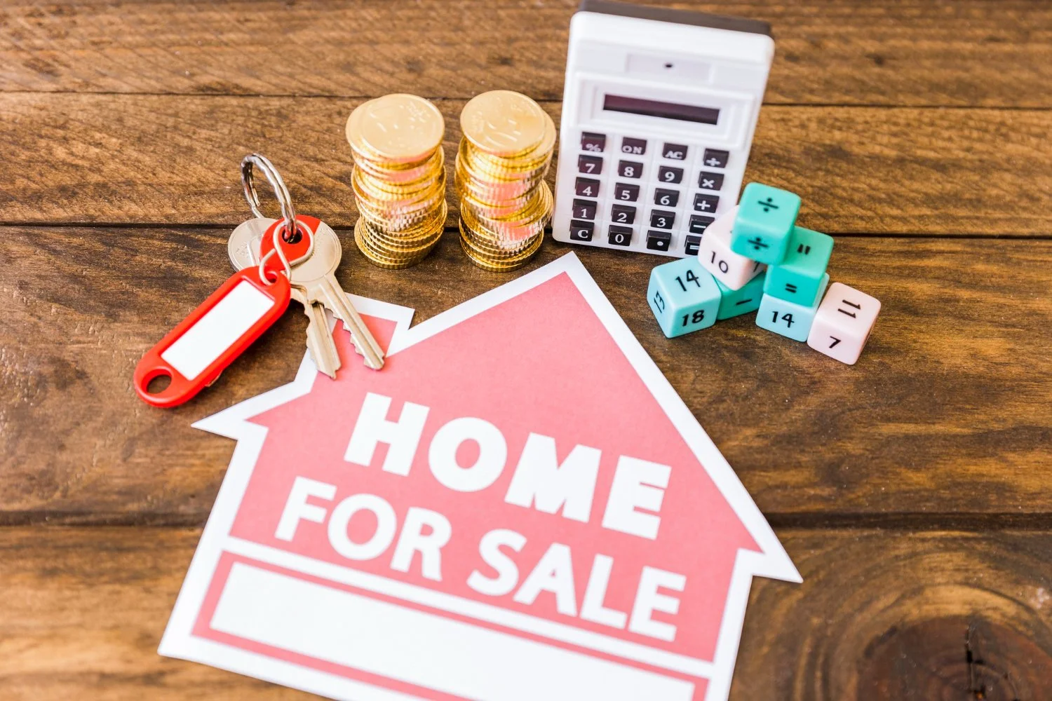 A 'Home for Sale' sign on a wooden surface surrounded by a keyring with keys, stacks of coins, a calculator, and colorful number and math operation cubes.