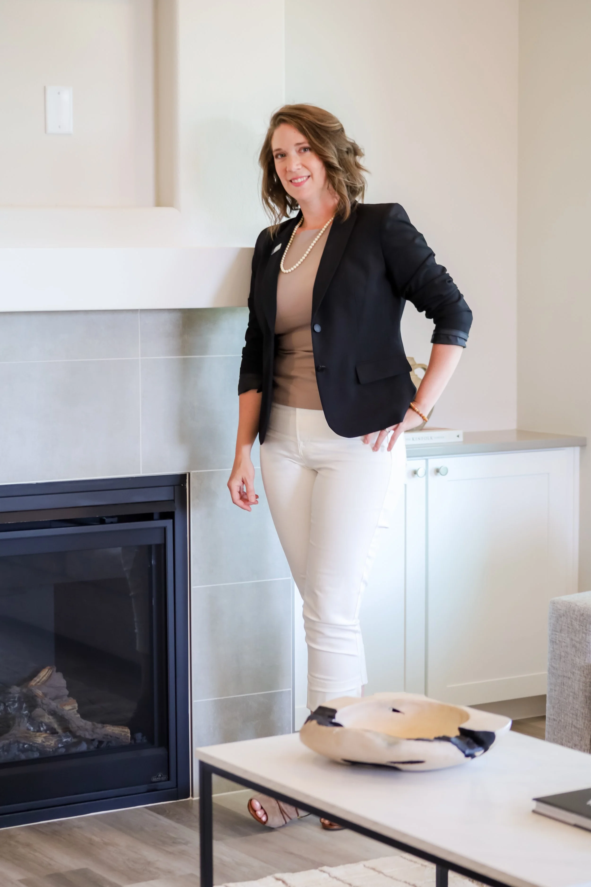 A woman with wavy hair wearing a black blazer, beige top, white pants, and a pearl necklace standing indoors near a fireplace and a dresser.