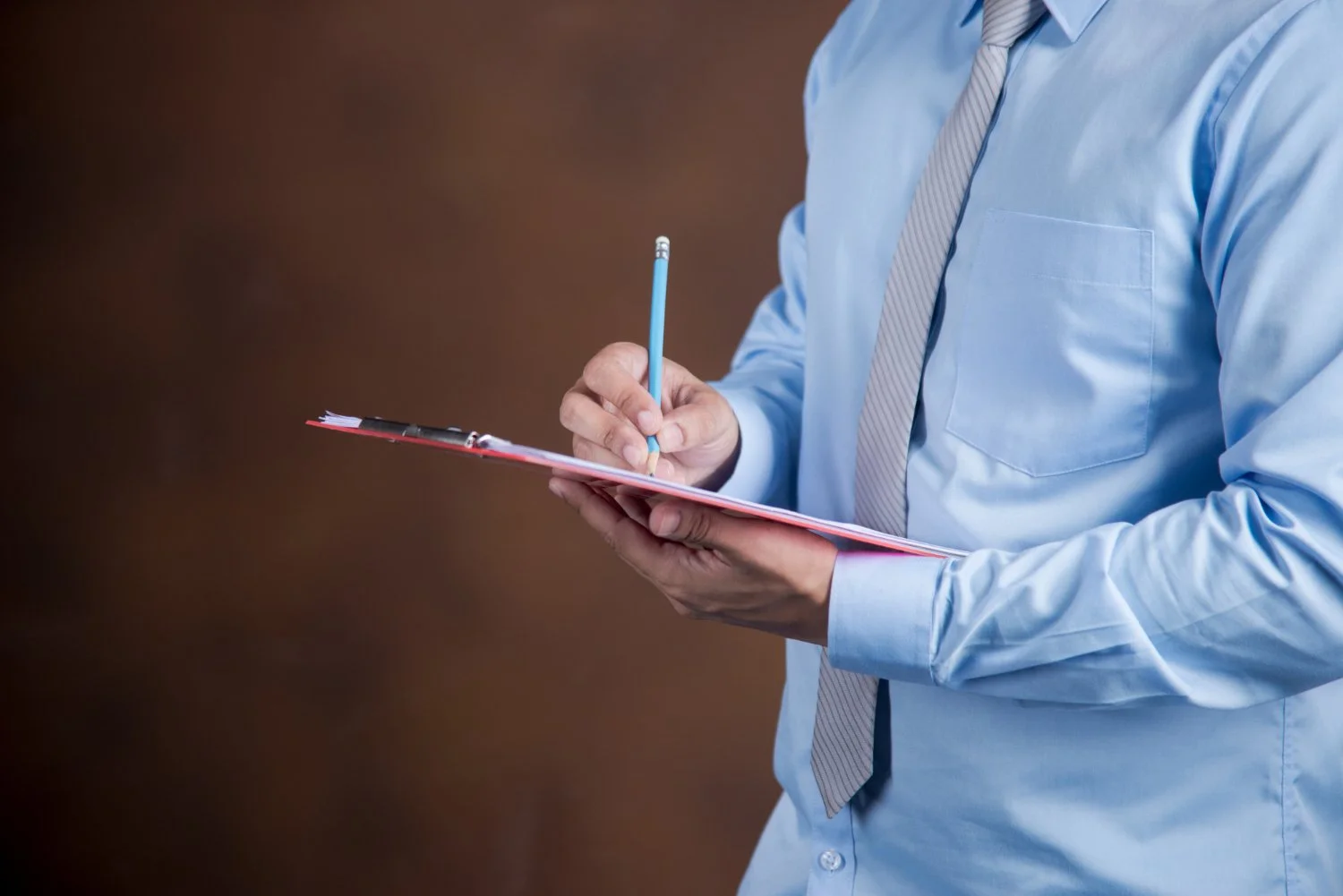 A man wearing a light blue dress shirt and gray striped tie writing on a clipboard with a blue pencil.