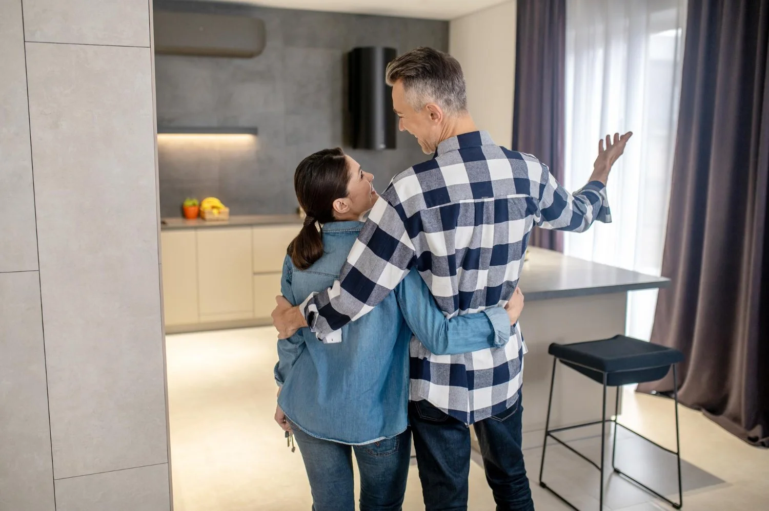 A couple happily embracing in a modern kitchen, with the man wearing a plaid shirt and the woman in a denim jacket, near a window with curtains.