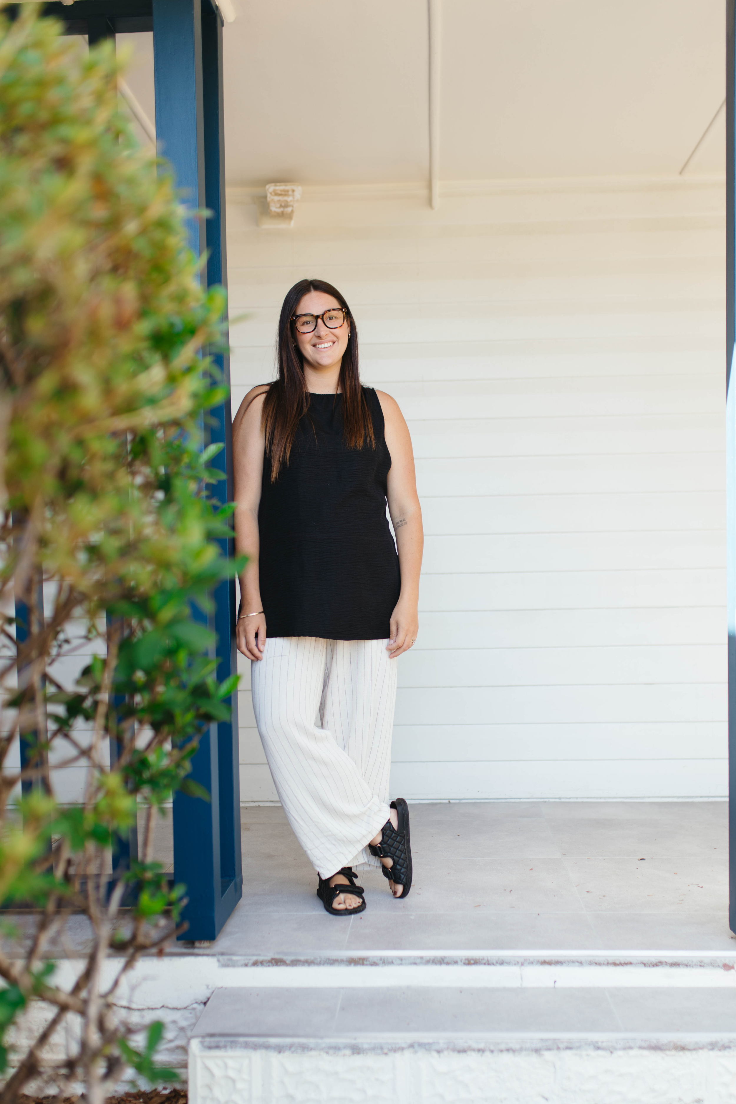 A woman with long dark hair, glasses, wearing a sleeveless black top, white striped pants, and black sandals, standing in the doorway of a building, smiling at the camera.