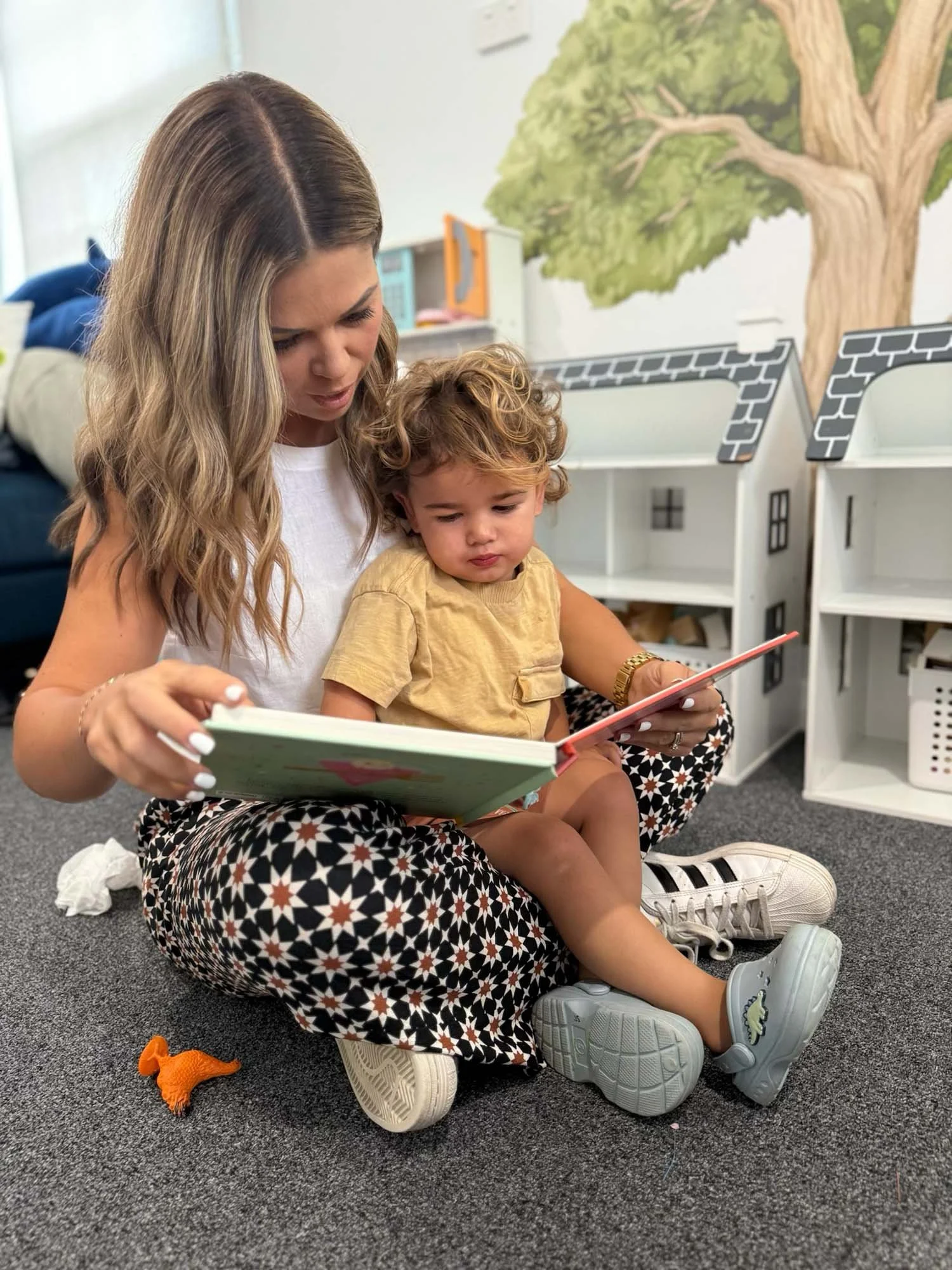 A woman and young child sitting on the floor reading a book together in a playroom with a mural of a tree on the wall.
