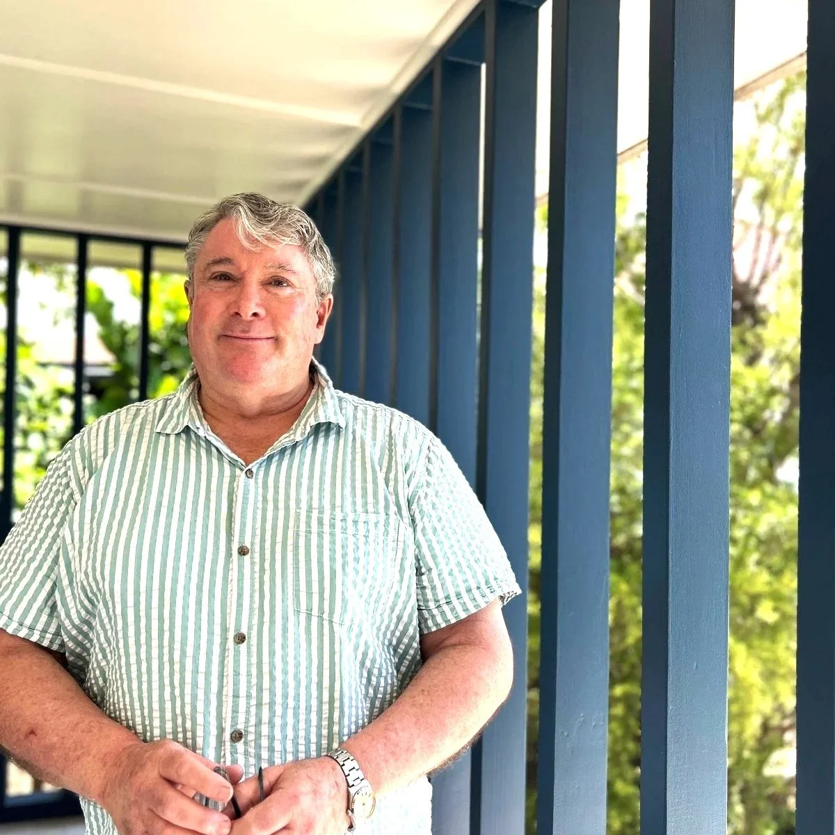A man standing on a balcony with blue railings and a background of trees and greenery, wearing a striped shirt and smiling.