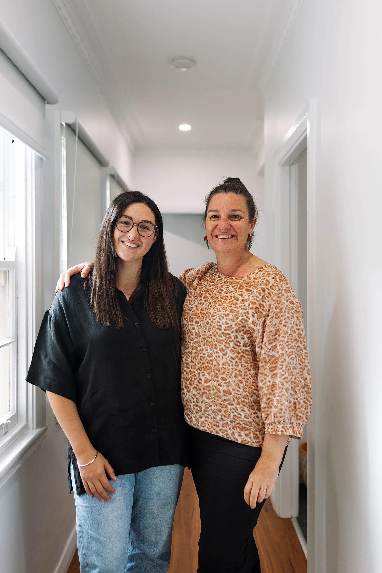 Two women smiling and standing close together in a hallway, one with her arm around the other.