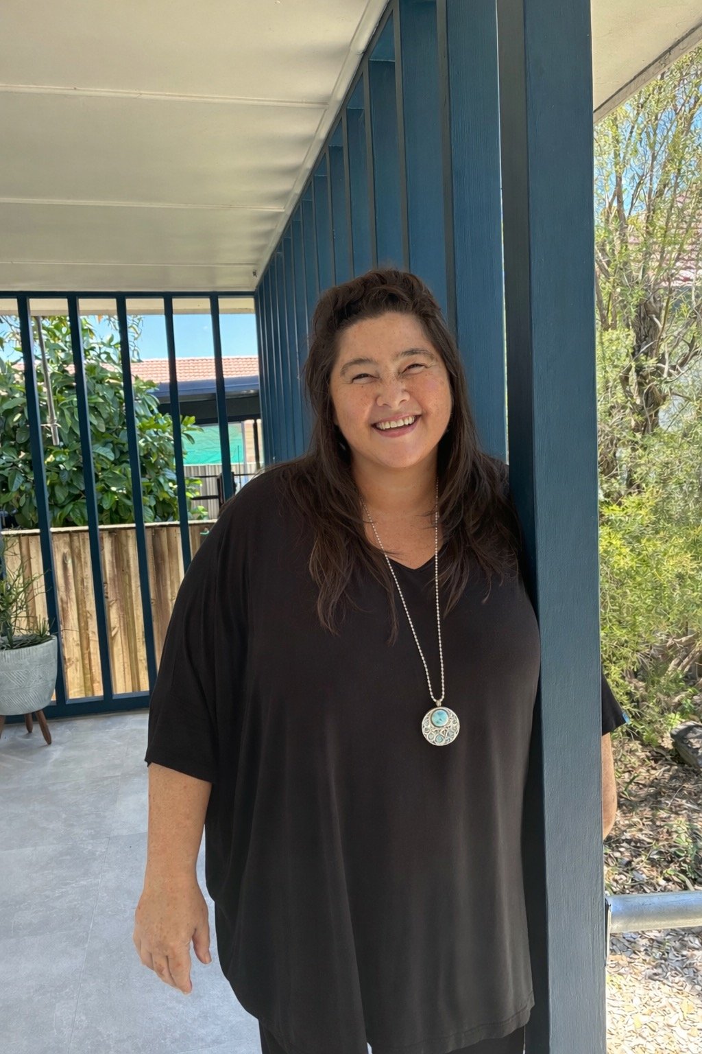 A woman smiling in a black shirt with a necklace, standing outdoors on a porch with blue vertical panels and greenery in the background.