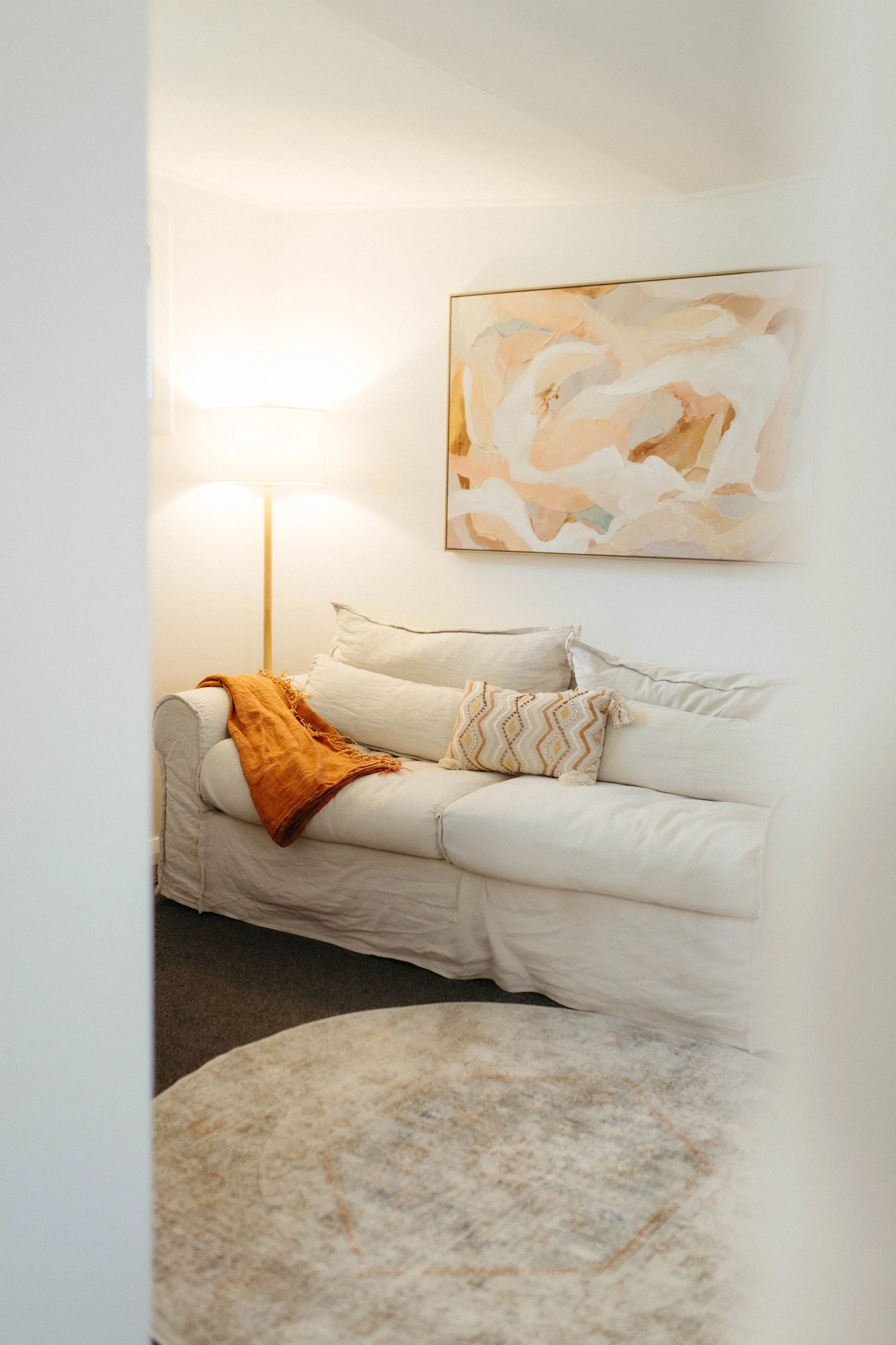 A cozy living room corner with a white sofa, a throw blanket, decorative pillows, a large wall art, a floor lamp, and a round rug.