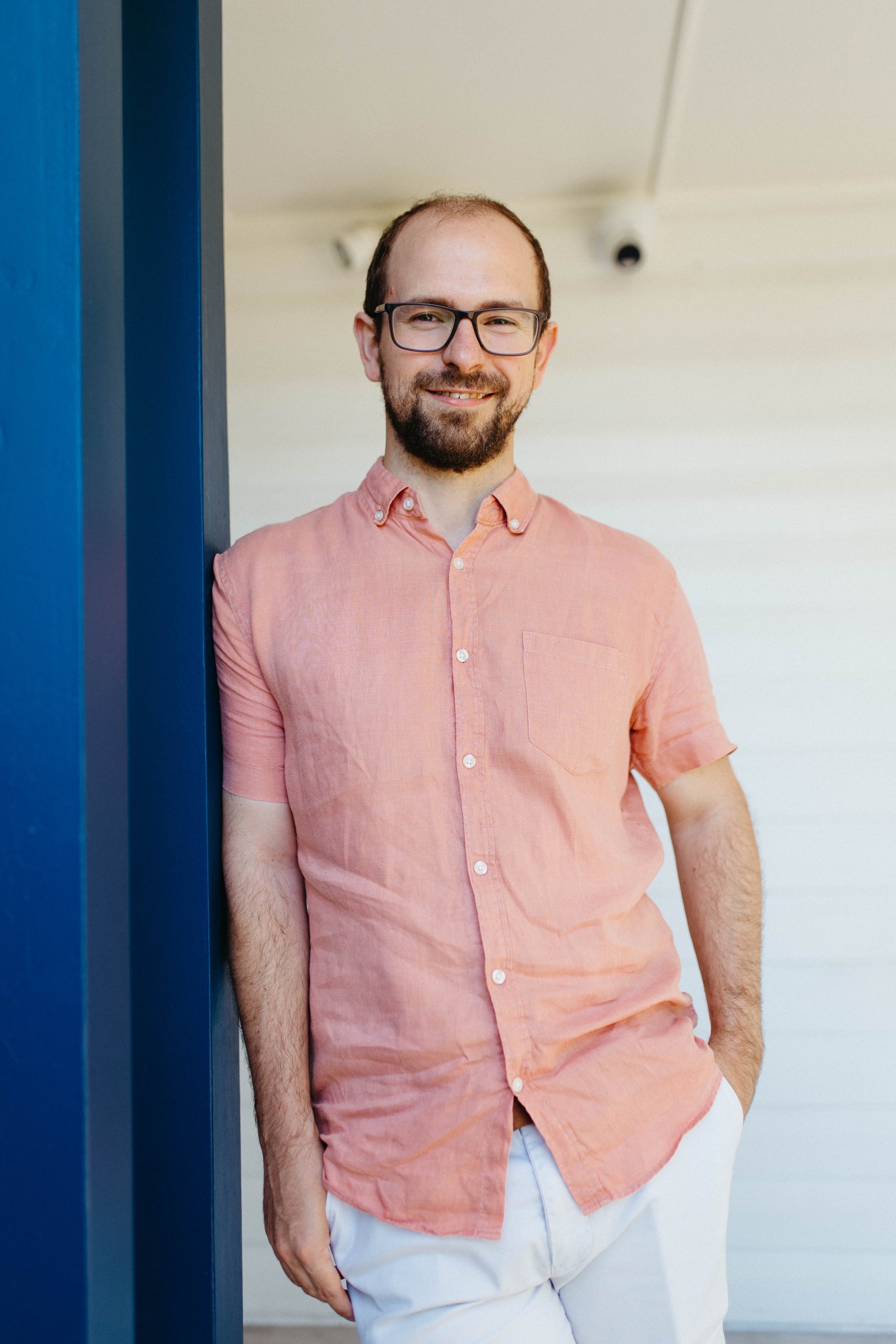 A man with glasses, a beard, and short hair, wearing a pink short-sleeve button-up shirt and white pants, standing outside leaning on a blue post with a white wall in the background and a security camera overhead.