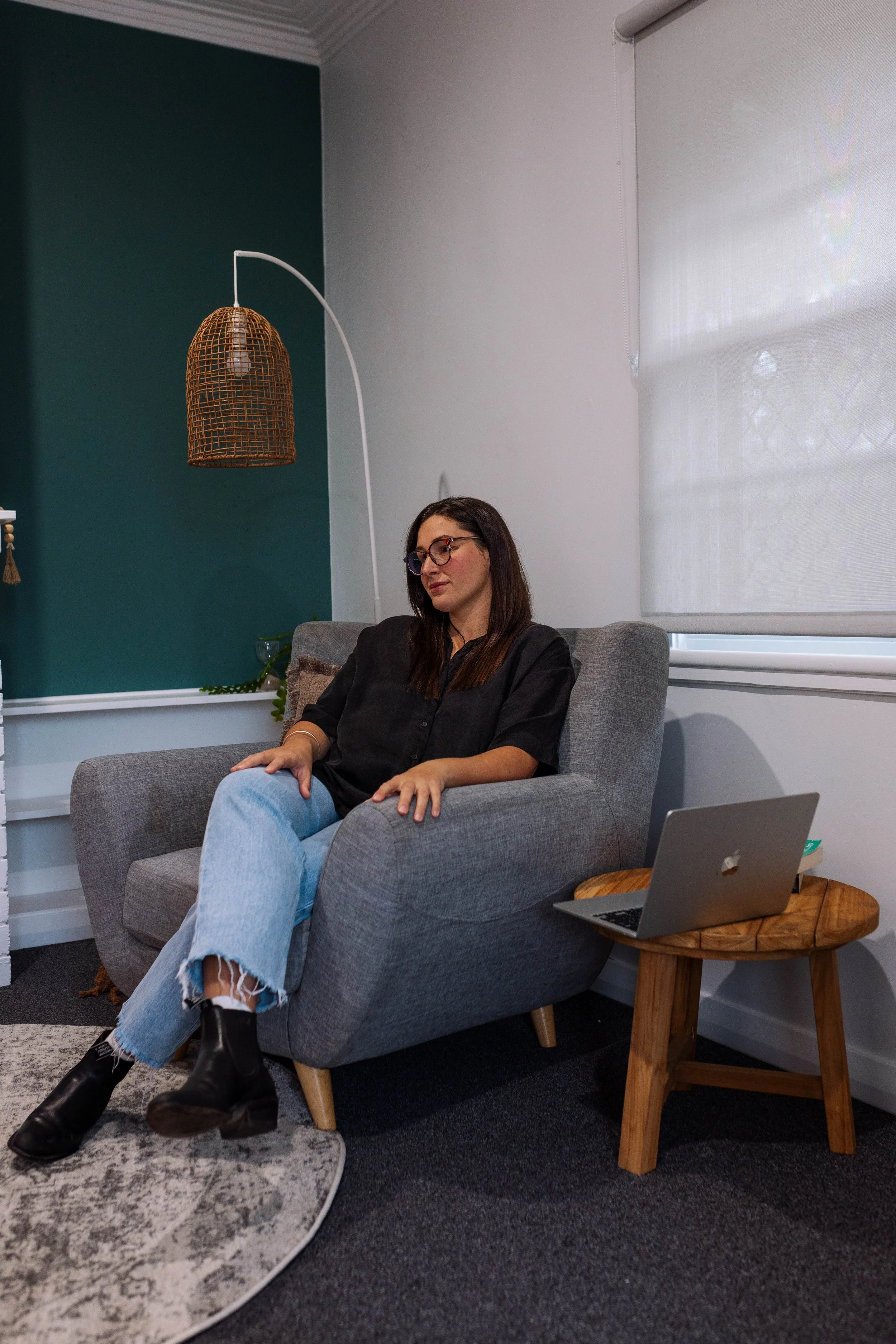 A woman with dark hair and glasses sitting in a gray armchair in a living room, next to a wooden side table holding an open silver laptop. There is a white window with a blind behind her and a woven hanging lamp above.