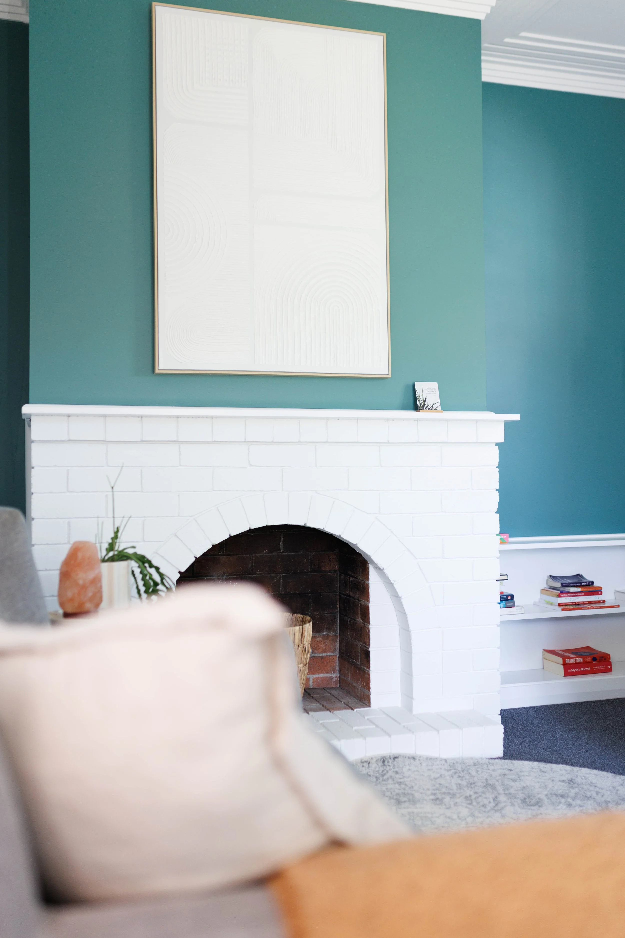 Living room with a white brick fireplace, teal painted wall above the fireplace, a large white and gold framed artwork on the wall, a small framed picture on the mantel, gray couch with a pillow, books on a shelves on the right side, and a salt lamp with a plant on a side table.