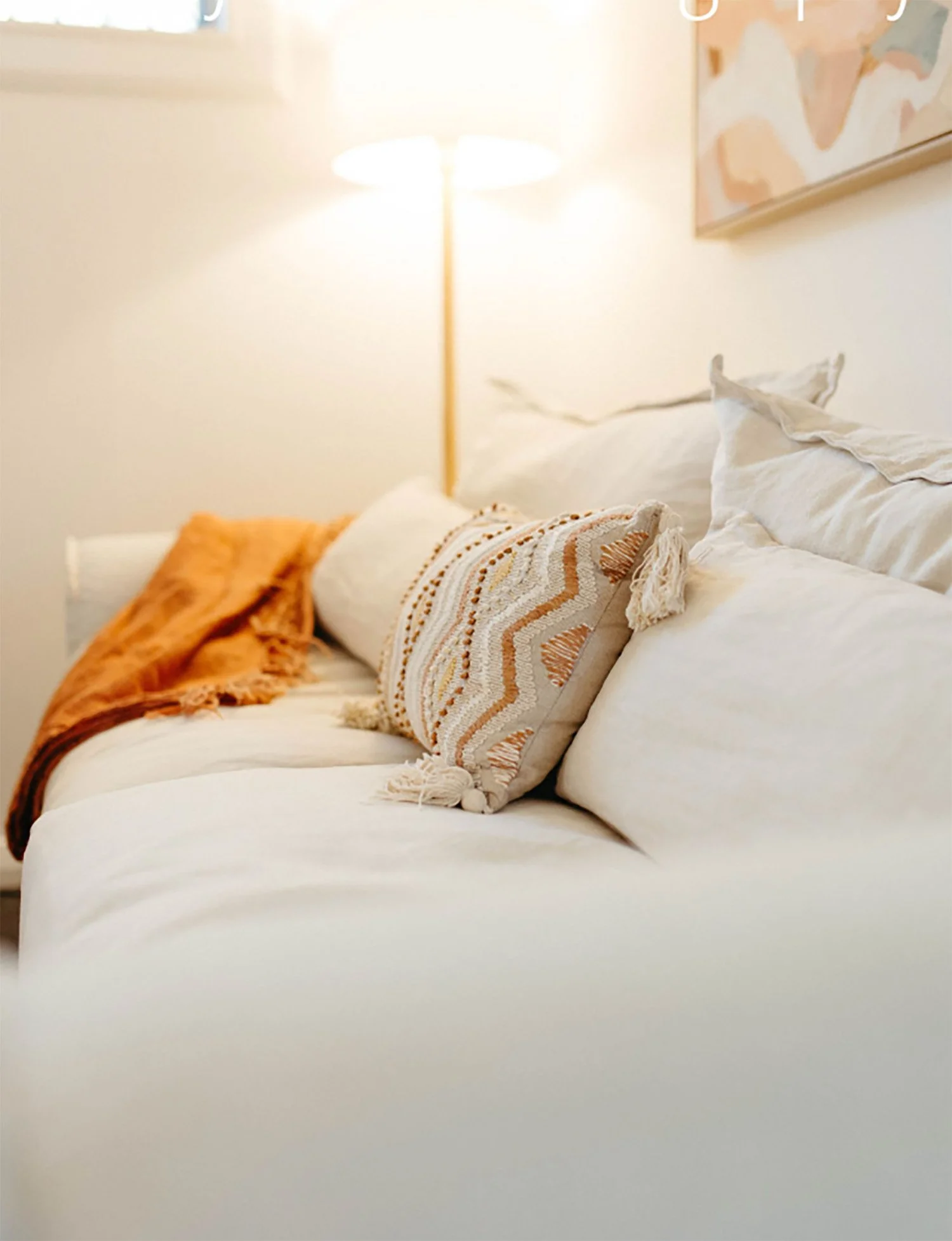 A cozy bedroom with a white bed, accented with decorative throw pillows and a rust-colored throw blanket. A soft lit lamp provides warm lighting, and a colorful abstract painting hangs on the wall.