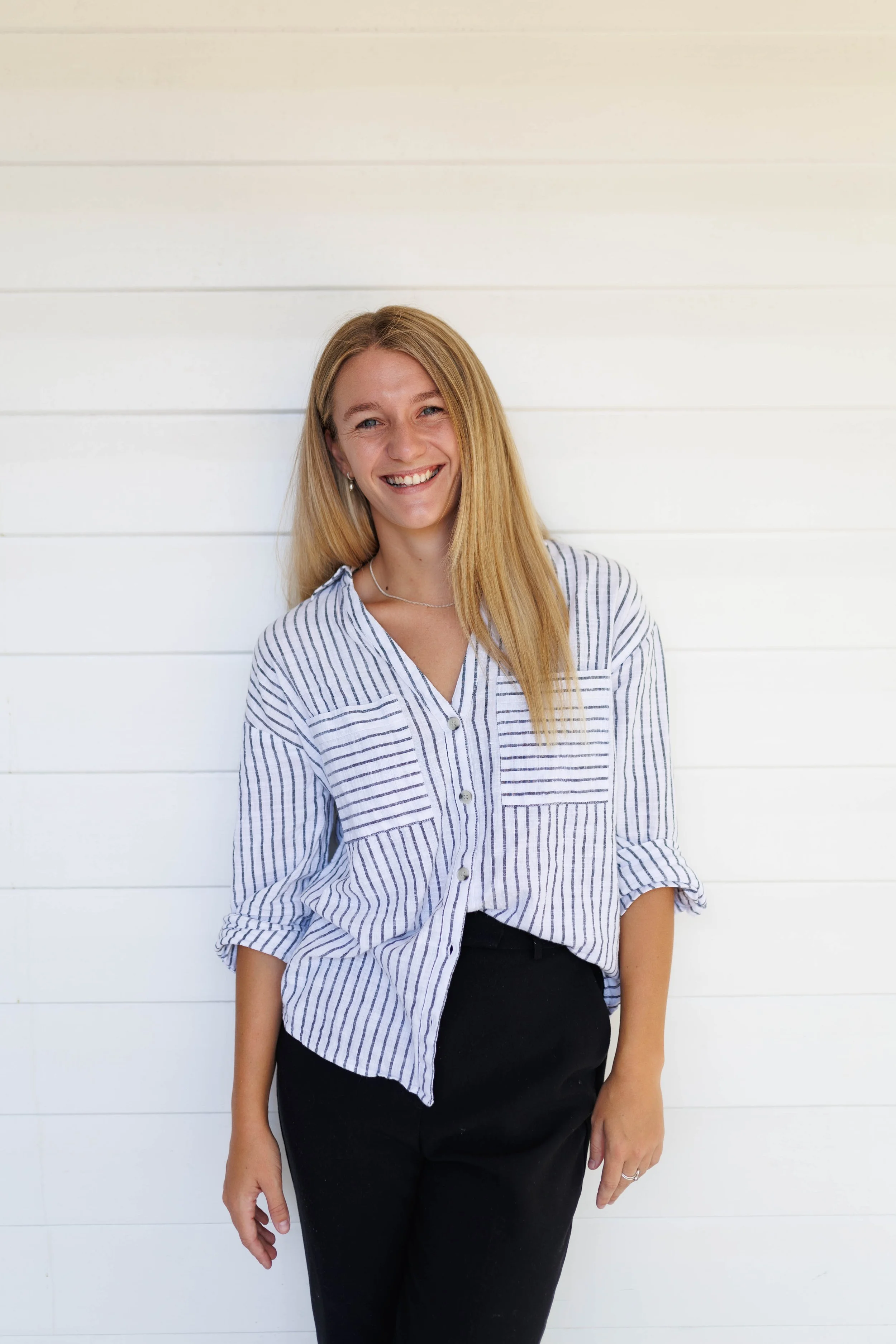 A woman with long blonde hair smiling, wearing a white and blue striped button-up shirt and black pants, standing against a white horizontal wooden wall.
