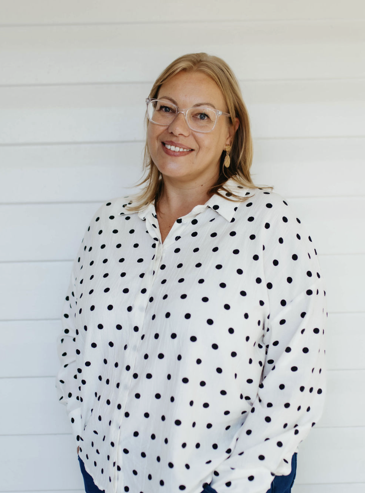 A woman with blonde hair, wearing glasses and a white blouse with black polka dots, standing in front of a white wooden wall, smiling at the camera.