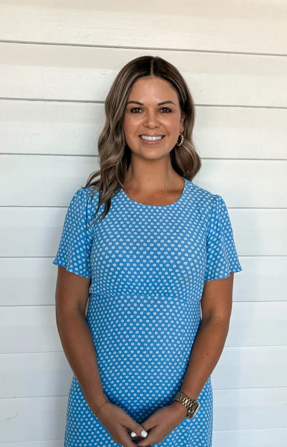A woman with shoulder-length brown hair and hoop earrings, wearing a blue dress with white polka dots, standing in front of a white horizontal wooden wall.