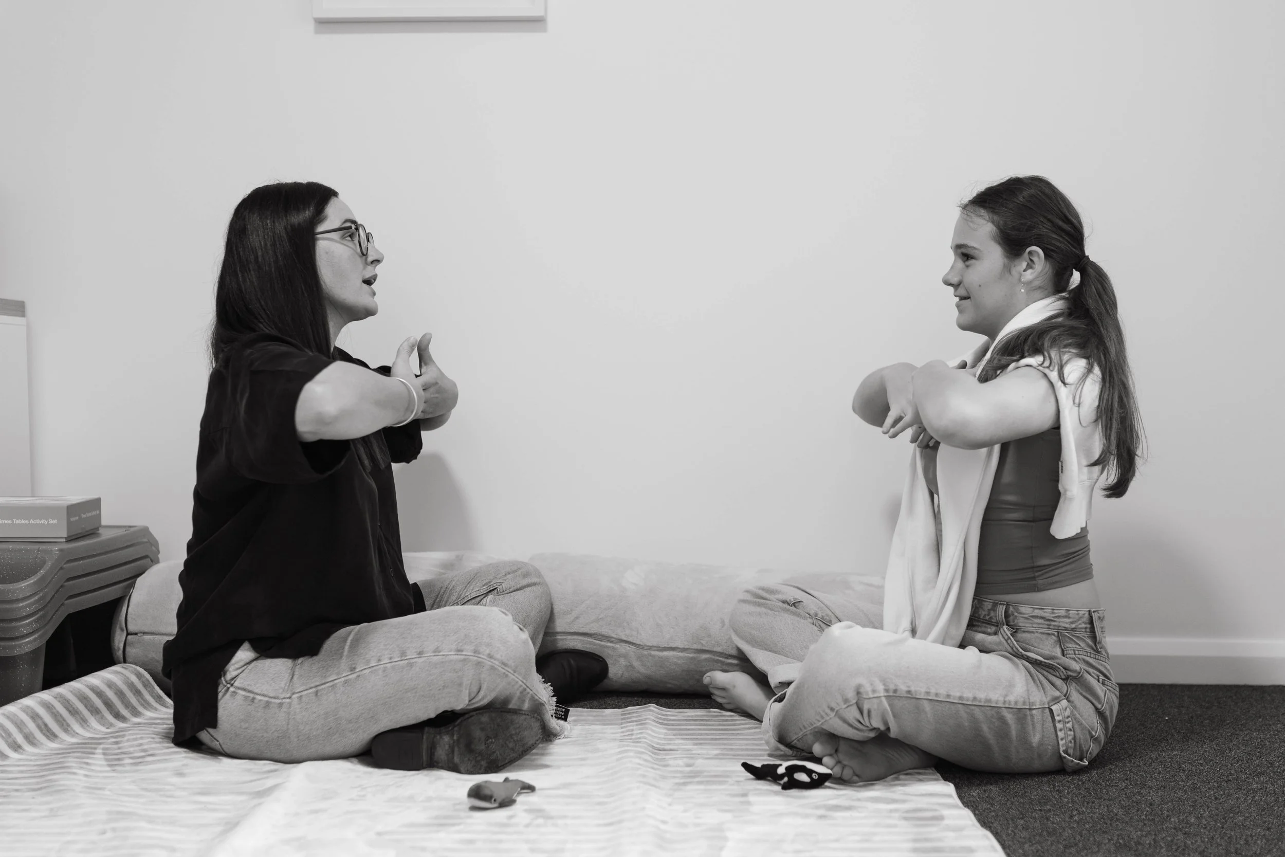 Two girls sitting cross-legged on a blanket, facing each other, engaging in a conversation or activity, in a room with a plain wall background.