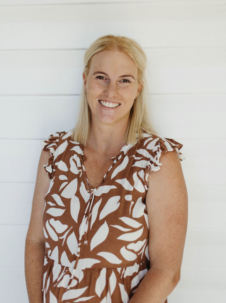 A woman with blonde hair, smiling, wearing a sleeveless brown dress with white leaf patterns, standing against a white paneled wall.