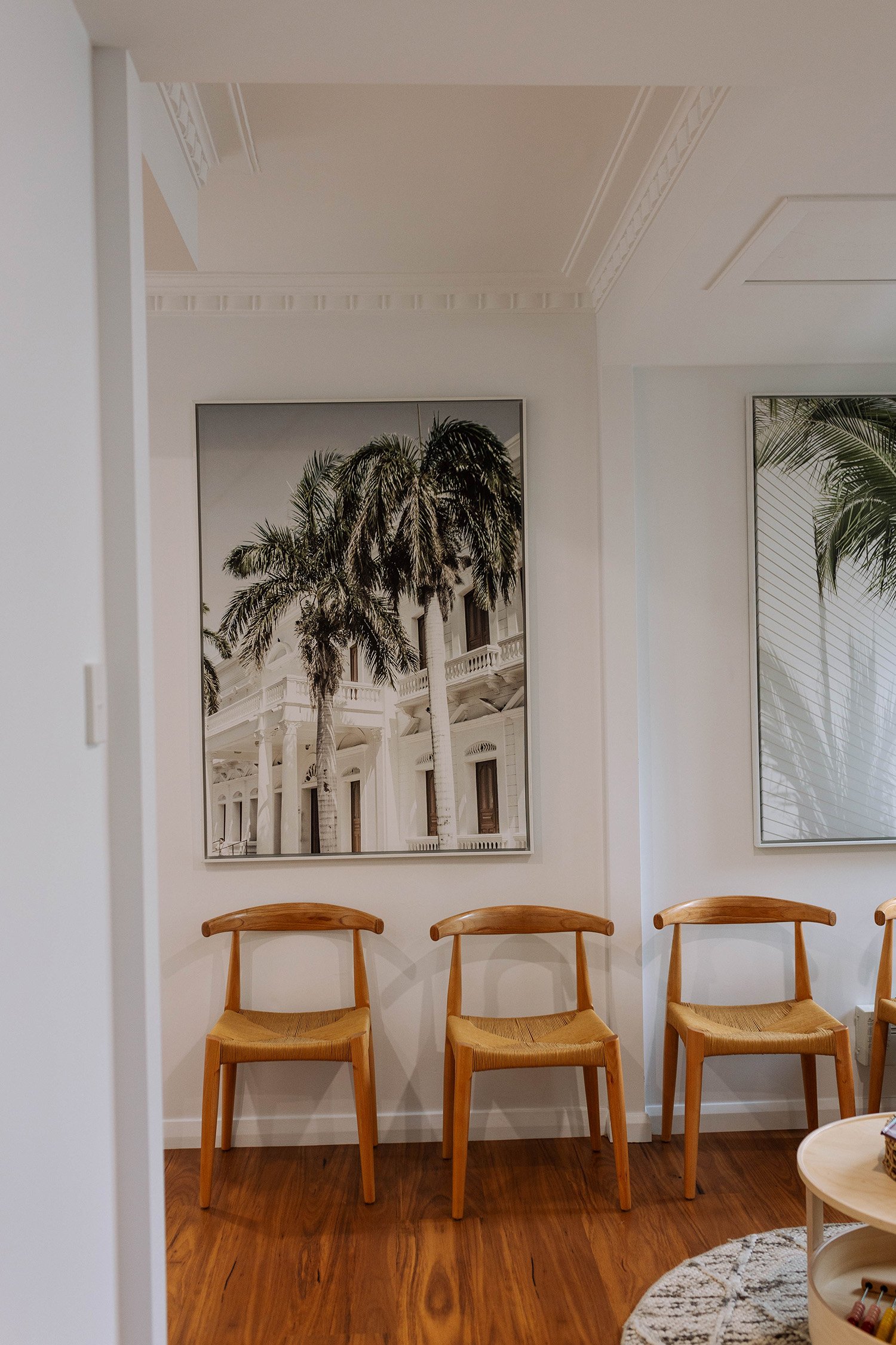 A waiting area with three wooden chairs with woven seats, a white wall with tropical houseplant artwork, and a hardwood floor.