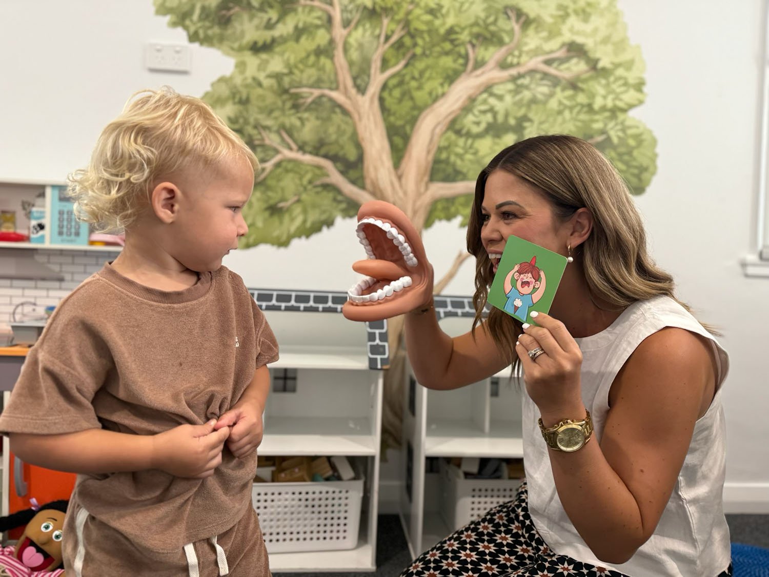 A young boy and a woman interact with a large mouth-shaped puppet. The woman holds a card with a cartoon girl and appears to be playfully pretending the puppet's mouth is the girl's voice. The setting is a colorful room with a tree mural on the wall.