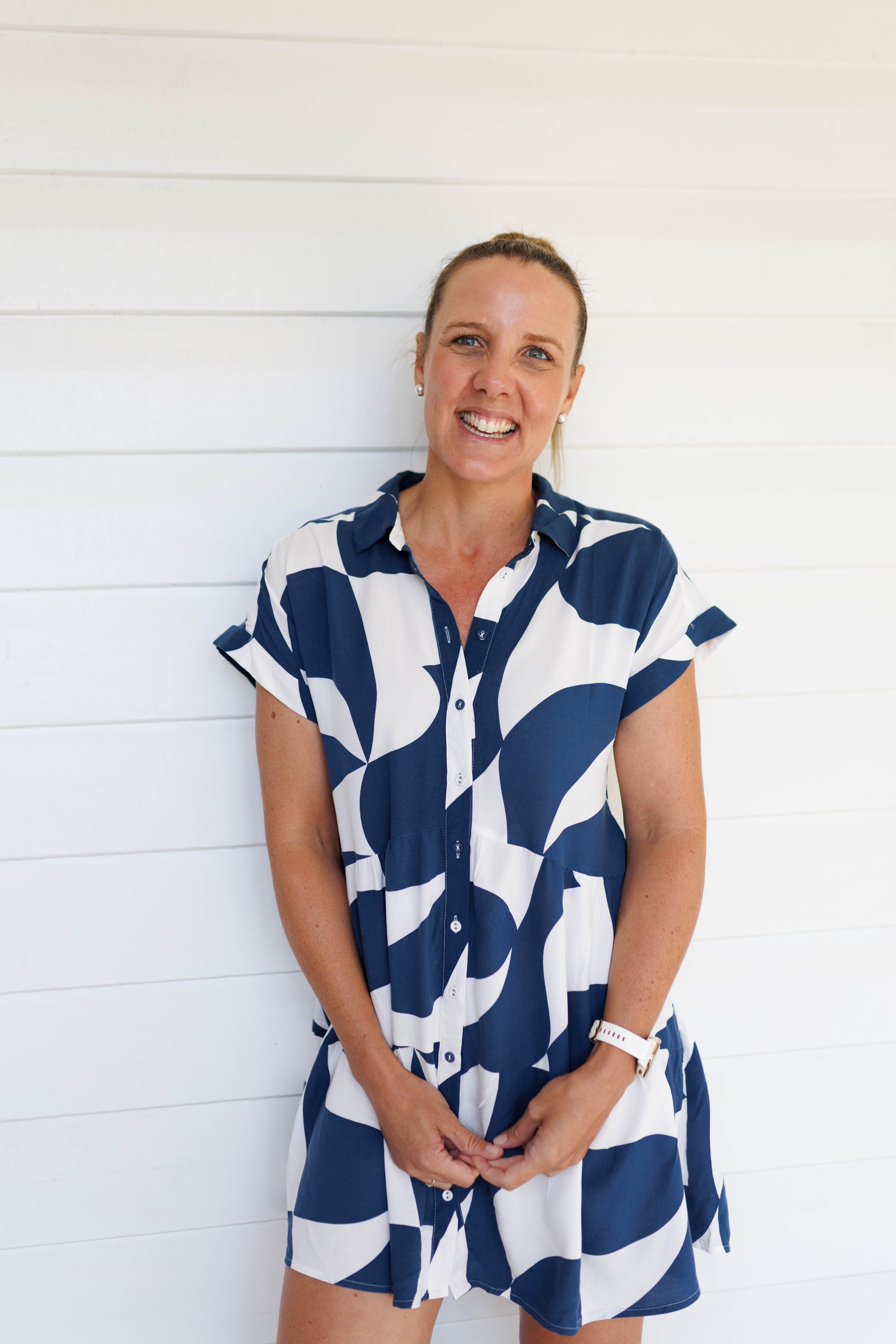 A woman with blonde hair tied back, smiling and wearing a blue and white patterned short-sleeve dress, standing against a white wooden panel wall.