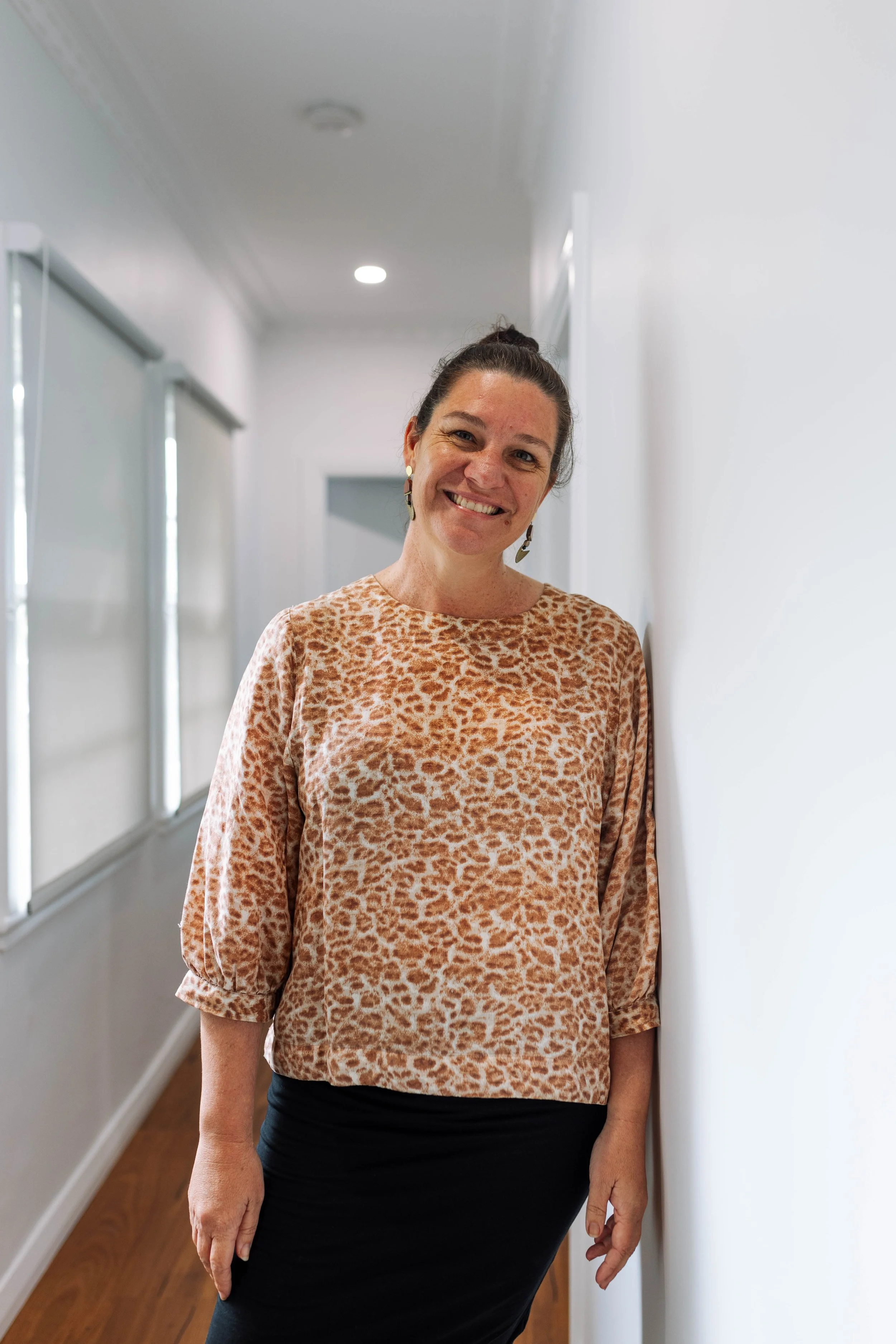 Smiling woman leaning against a white wall in a hallway, wearing a leopard print blouse and a black skirt.
