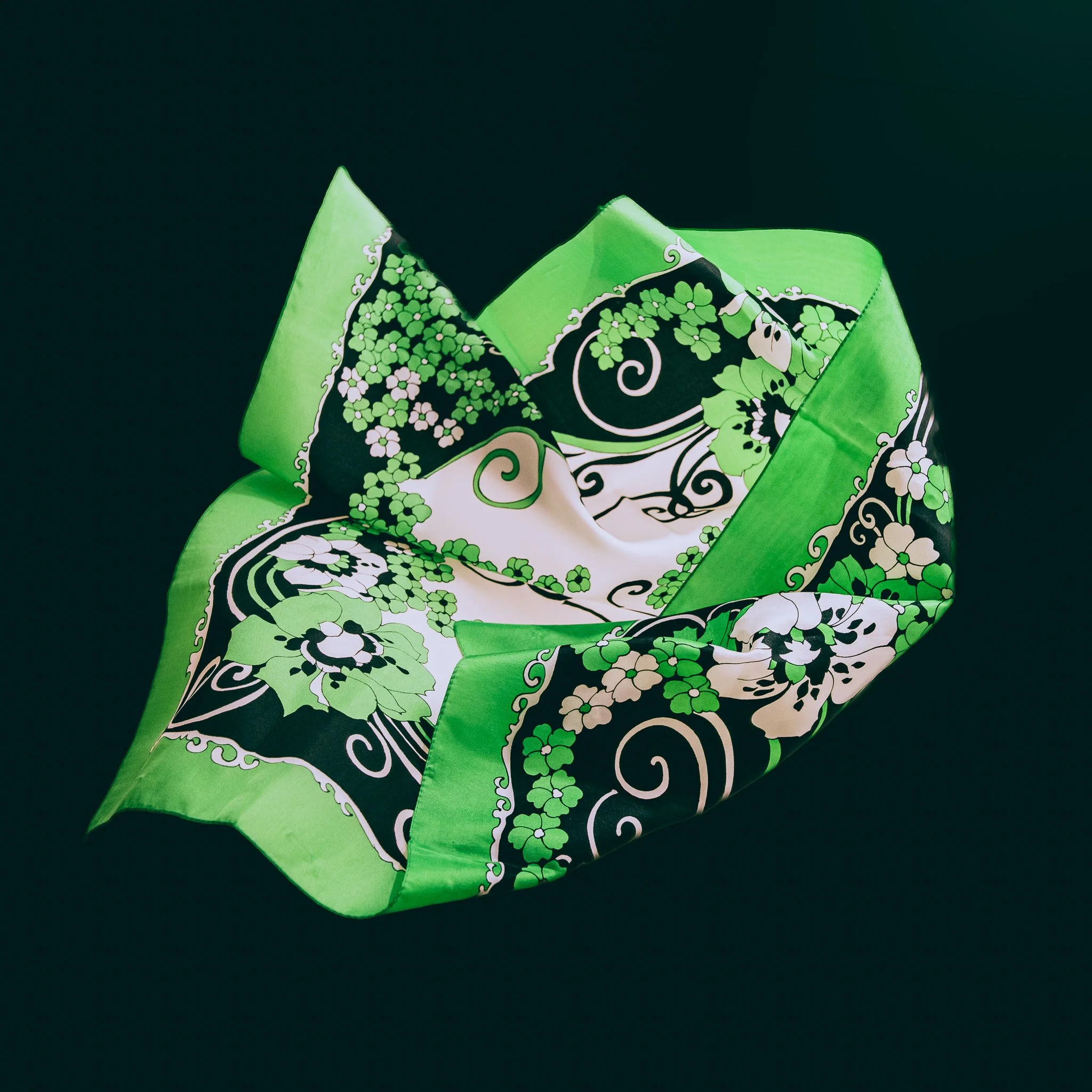 A silk scarf with a floral and vine pattern in black, white, and shades of green, arranged in a loose, swirling shape on a dark background.