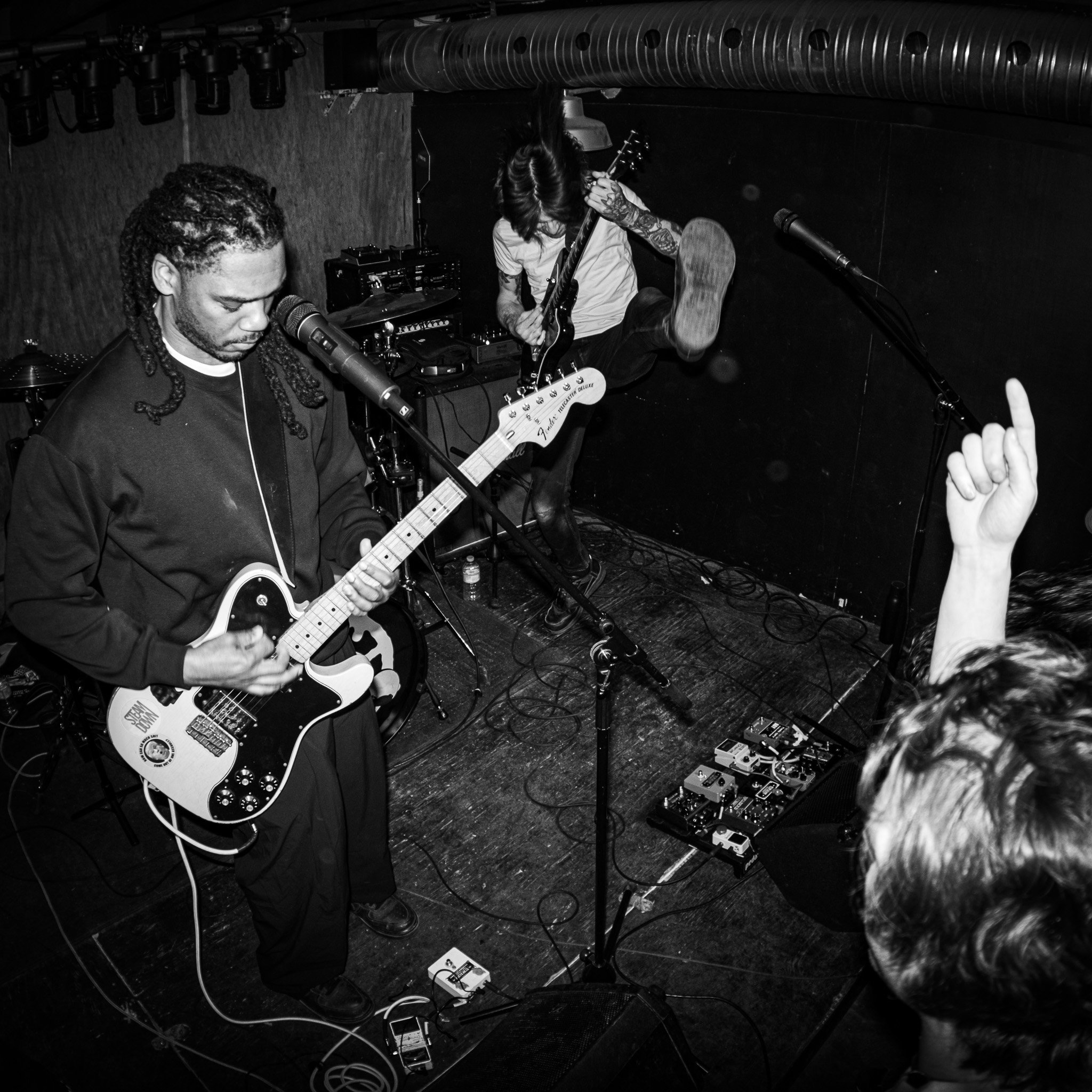 Two musicians performing on stage in a small venue. One is playing an electric guitar, and the other is jumping in the background with a guitar. A person in the audience is raising a finger. The scene is black and white.