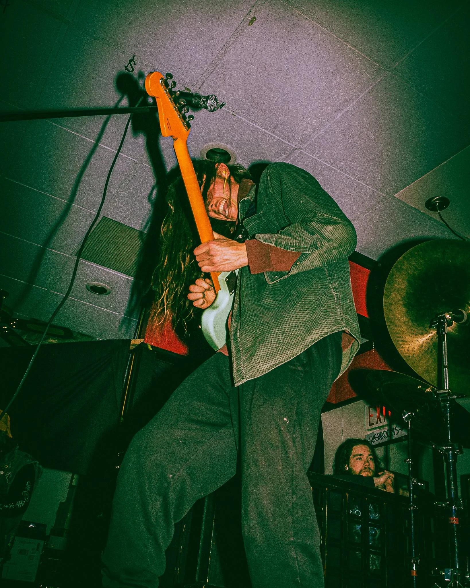 A person with long hair is playing an electric guitar on stage, leaning back and smiling, with drum equipment visible in the background.