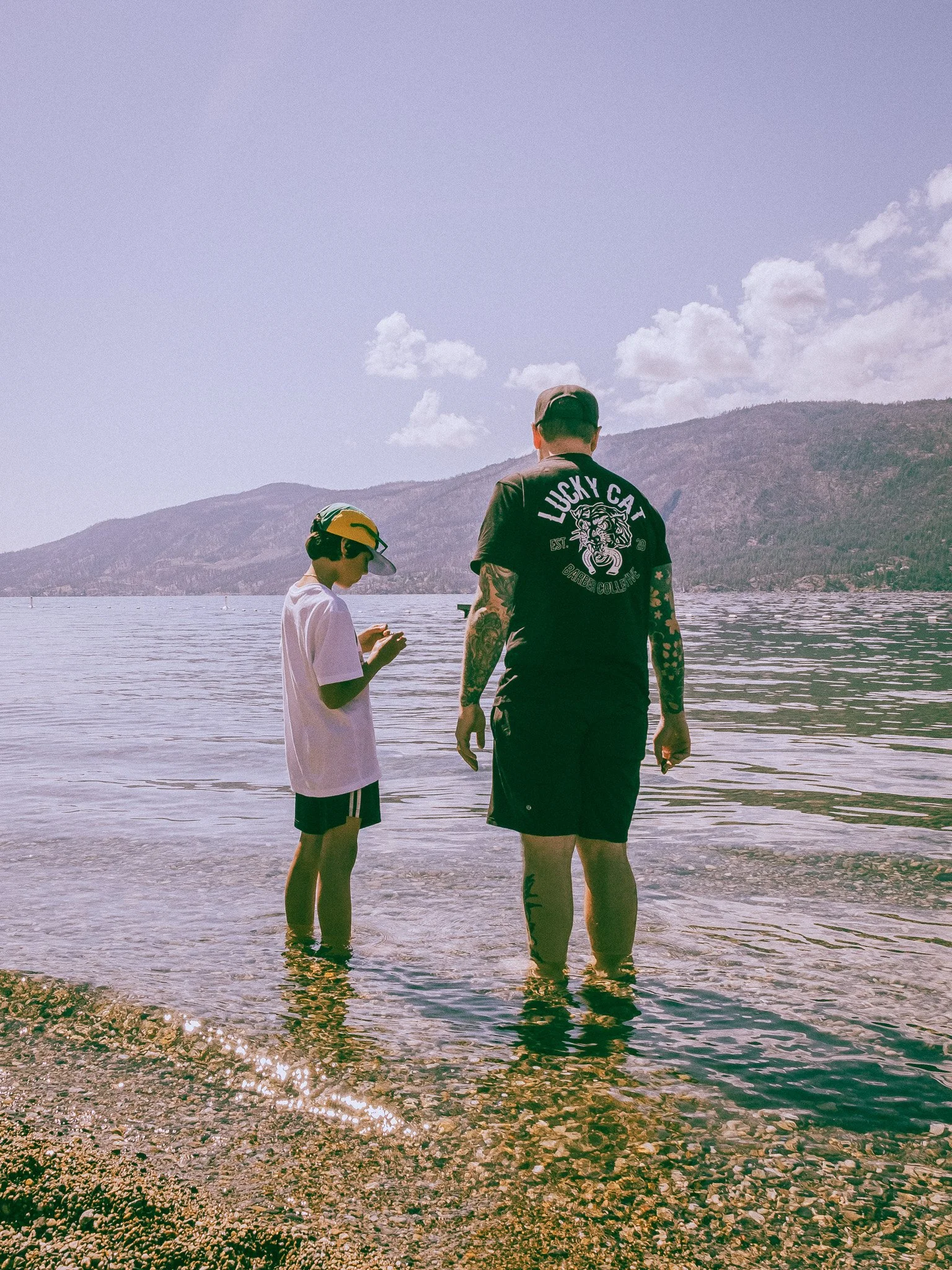 Two people standing in shallow water on a beach, with mountains and a cloudy sky in the background. One is a child wearing a cap, t-shirt, and shorts, looking at something in their hands; the other is an adult with tattoos wearing a black t-shirt and