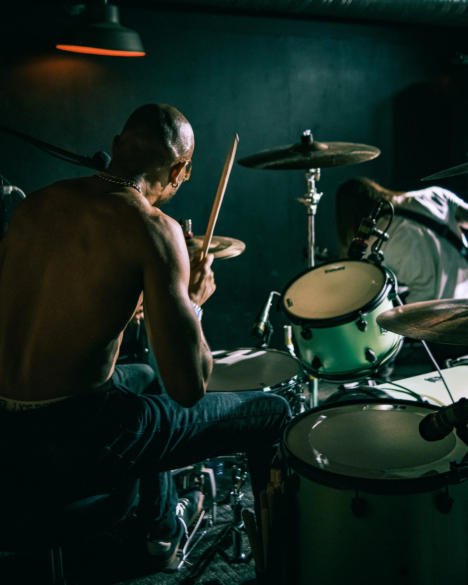 A shirtless drummer playing drums in a dark room with minimal lighting.