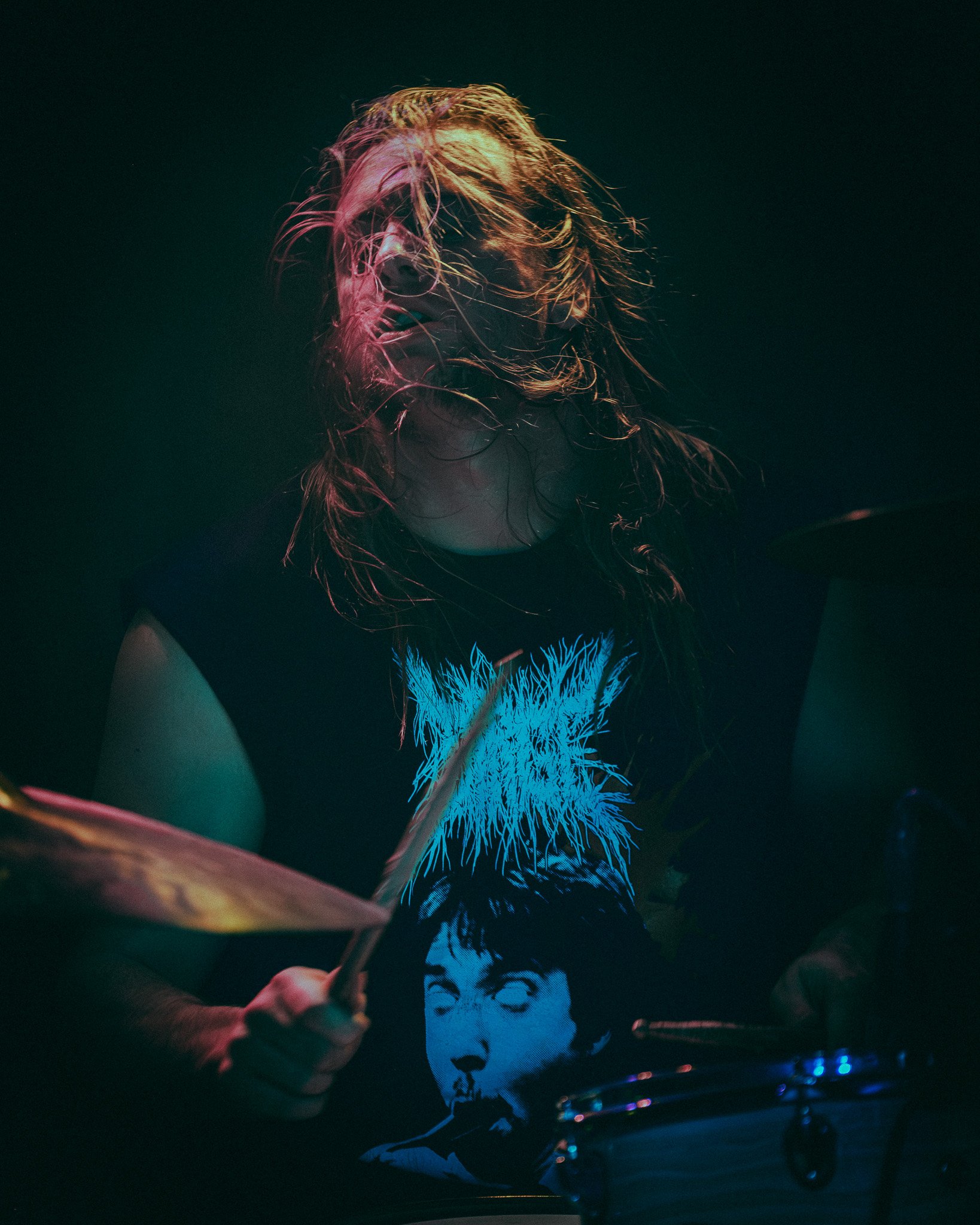 A musician with long hair playing drums in a dark setting, wearing a black t-shirt with a graphic of a face and neon-colored artwork.