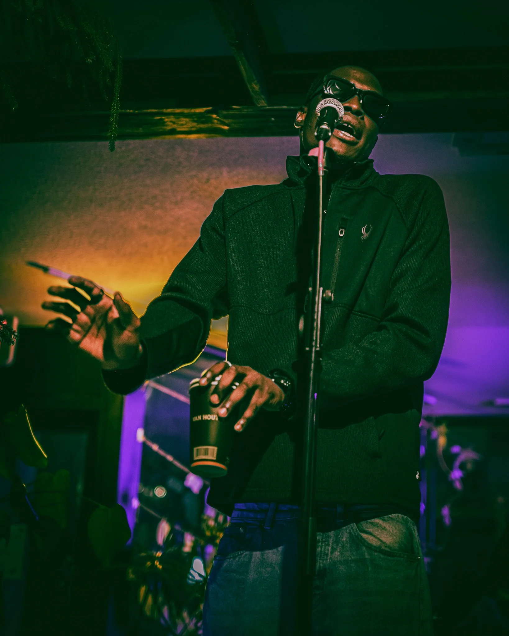 A man wearing sunglasses and a black jacket standing in front of a microphone on stage, holding a cup, with colorful stage lighting in the background.