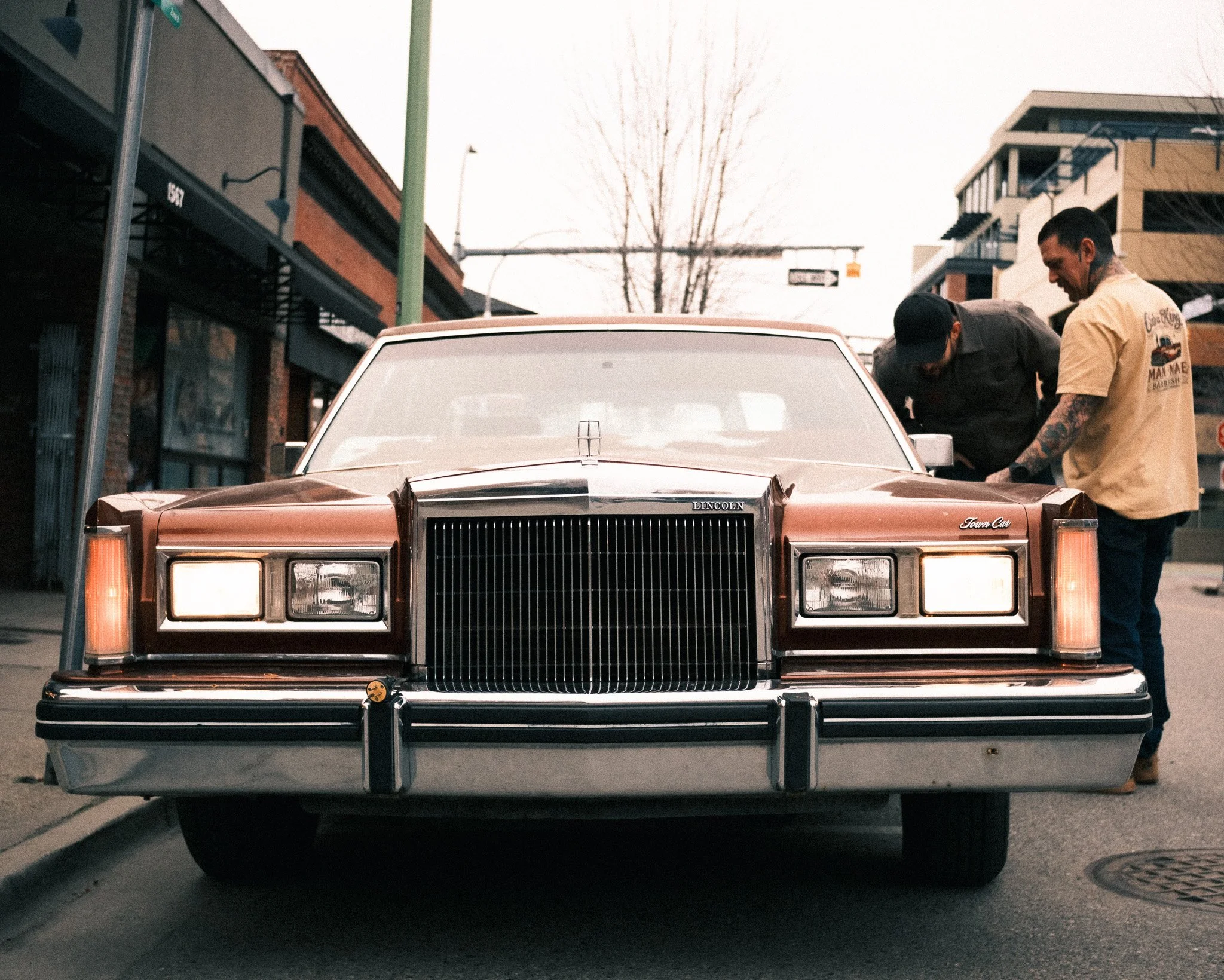 A vintage brown Lincoln Town Car parked on the street with two men examining it, one wearing a beige shirt with tattoos and the other in black clothing, in an urban setting.