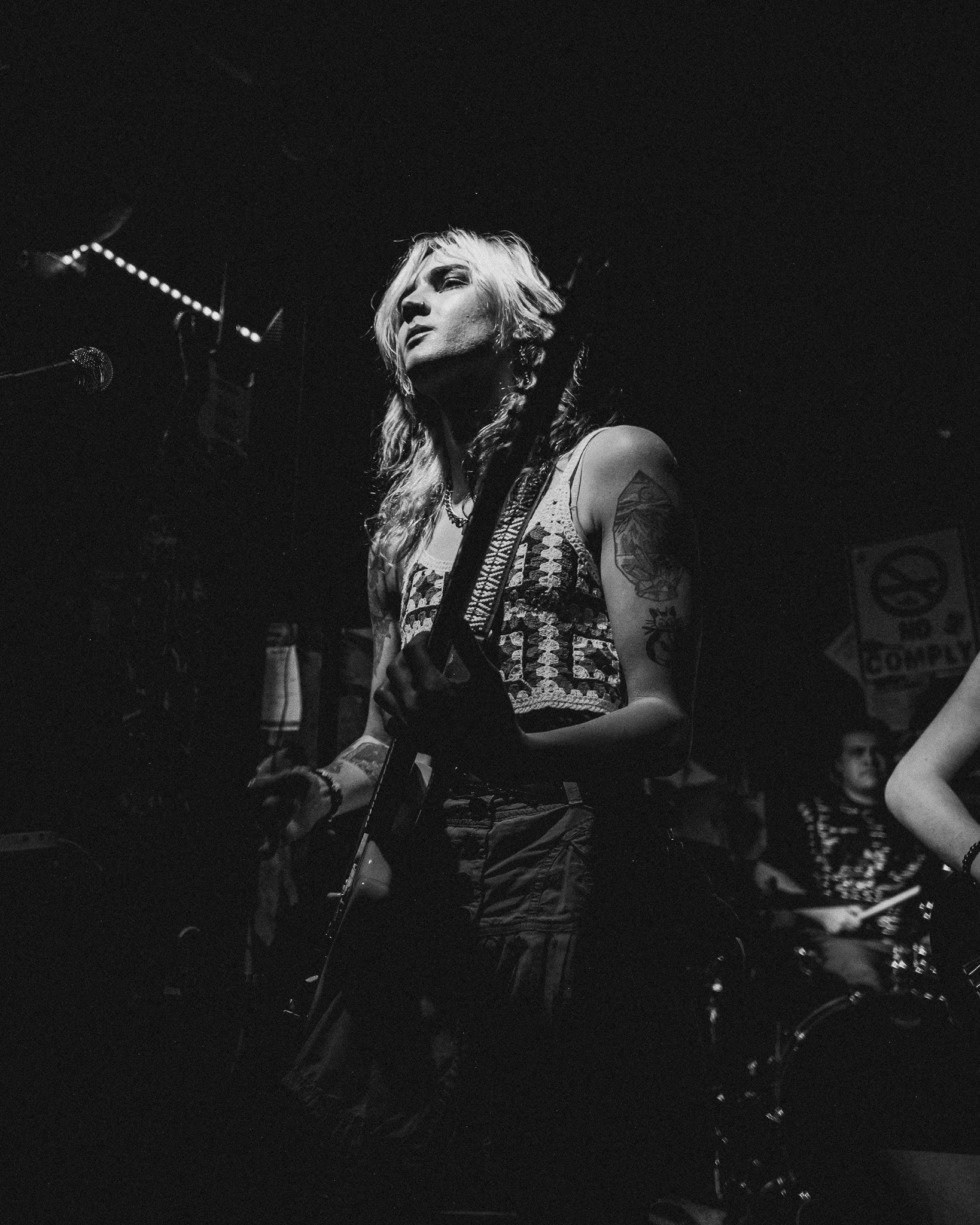 A black and white photo of a woman with long wavy hair, tattoos on her arms, wearing a sleeveless top, holding a guitar on stage.