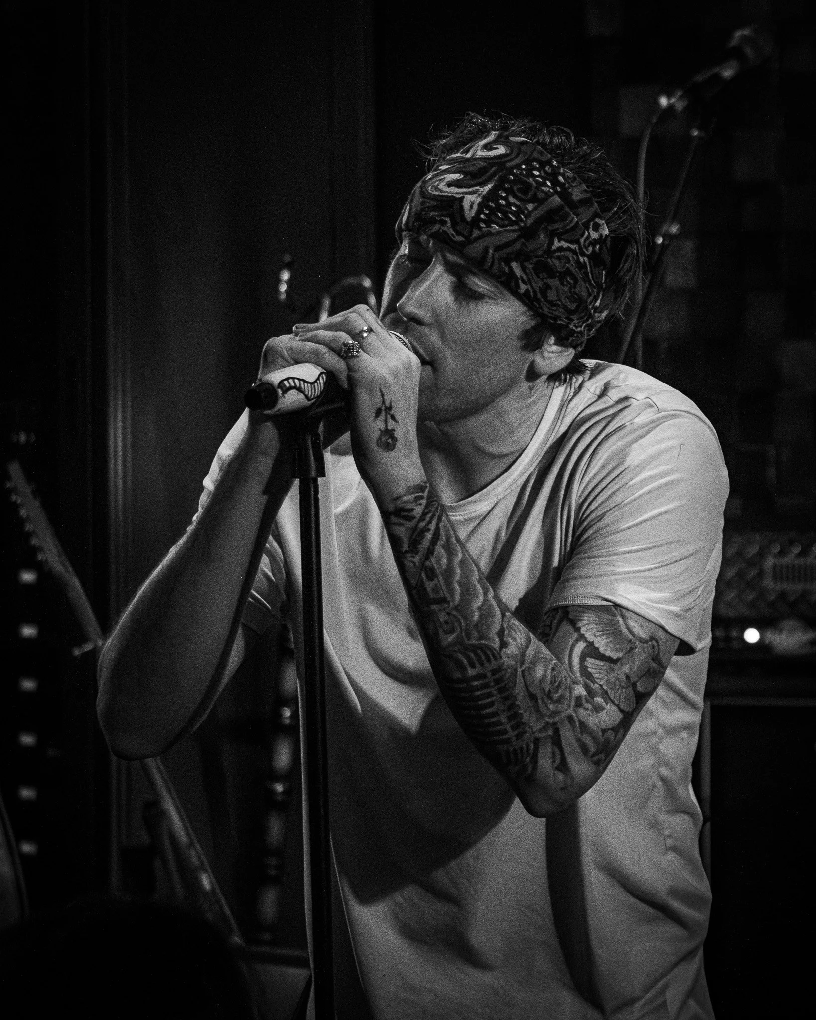 A male singer with tattoos on his left arm, wearing a bandana on his head, singing into a microphone in a recording studio.