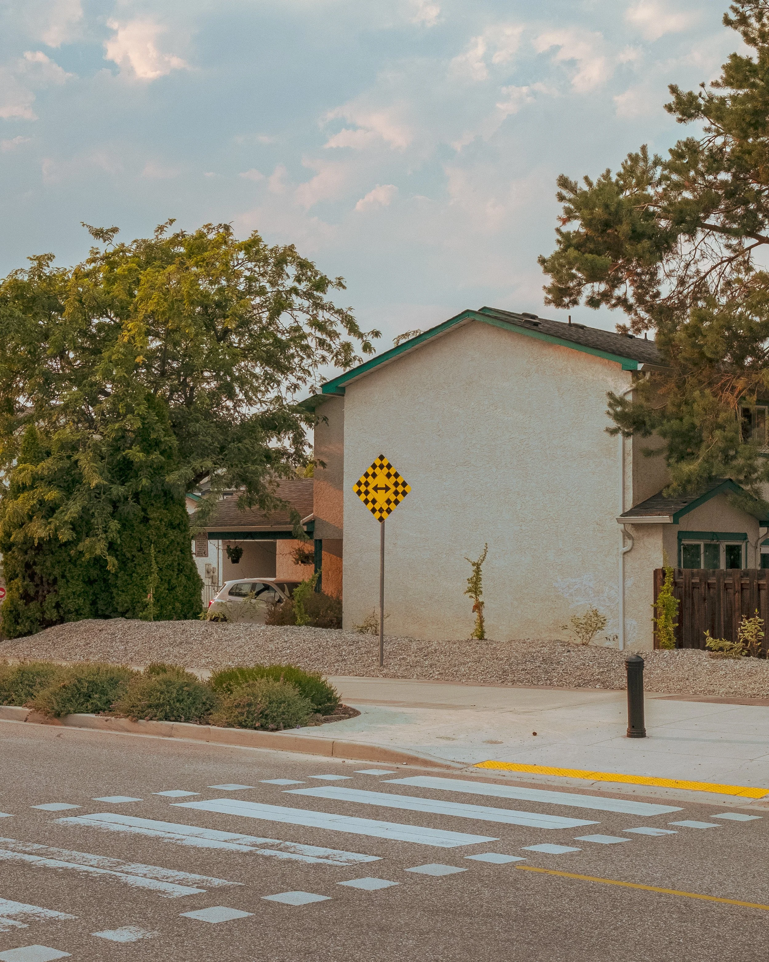 A suburban street corner with a pedestrian crosswalk, a curved yellow line, and a yellow warning sign indicating a merging traffic lane. There are trees, shrubs, and houses in the background under a partly cloudy sky.