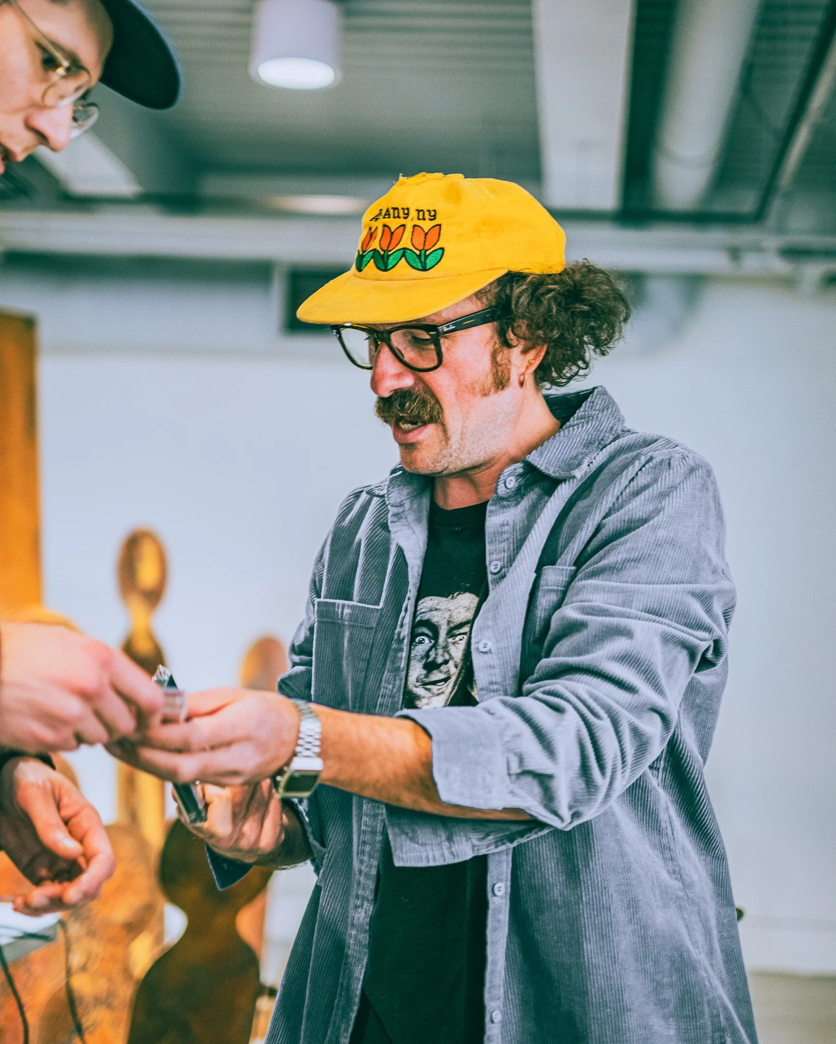 A man with curly hair, glasses, and a mustache wearing a yellow cap with tulip flowers and the text 'ANY NY', a black graphic T-shirt, and a gray striped button-up shirt. He is talking to another person whose hands are visible, exchanging items in a 