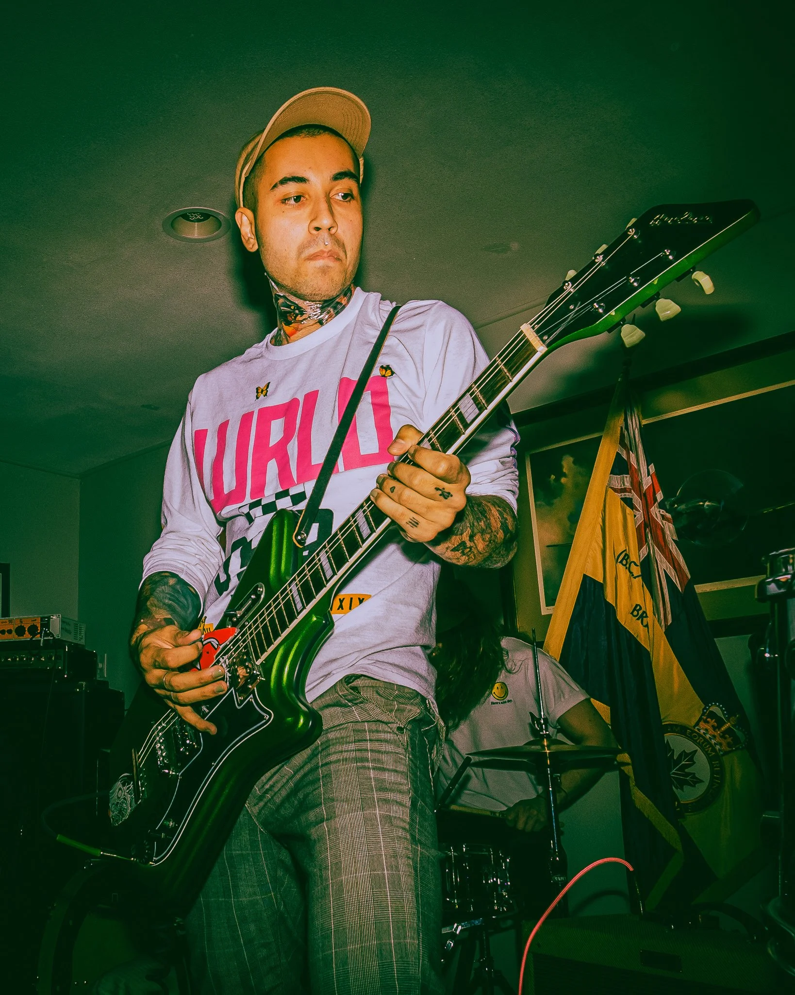 A man playing an electric guitar in a room with graffiti-style decor and flags, wearing a beige cap, a white shirt with large pink text, and plaid pants.