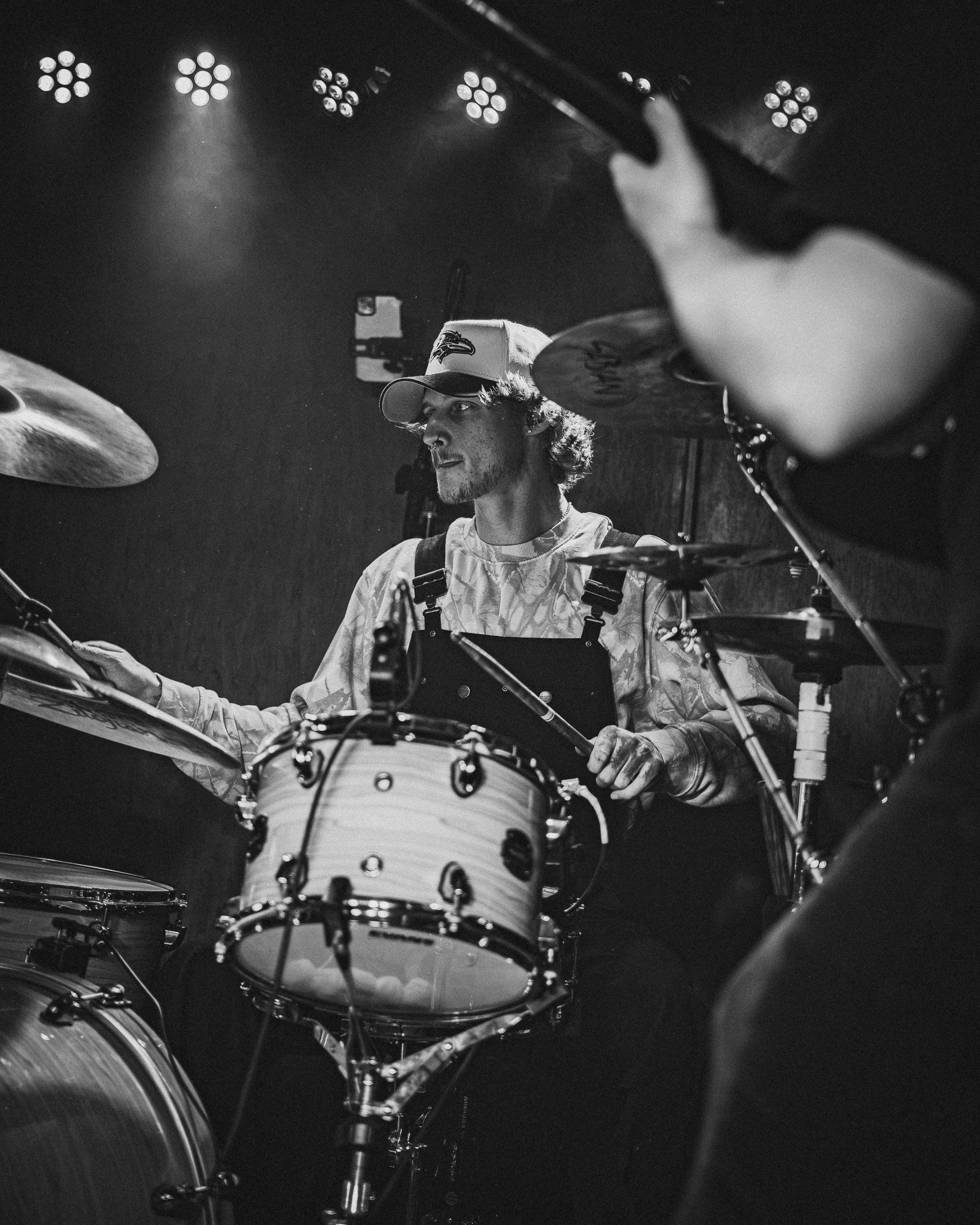A musician playing drums during a performance, wearing a baseball cap and casual clothing, with stage lights overhead.