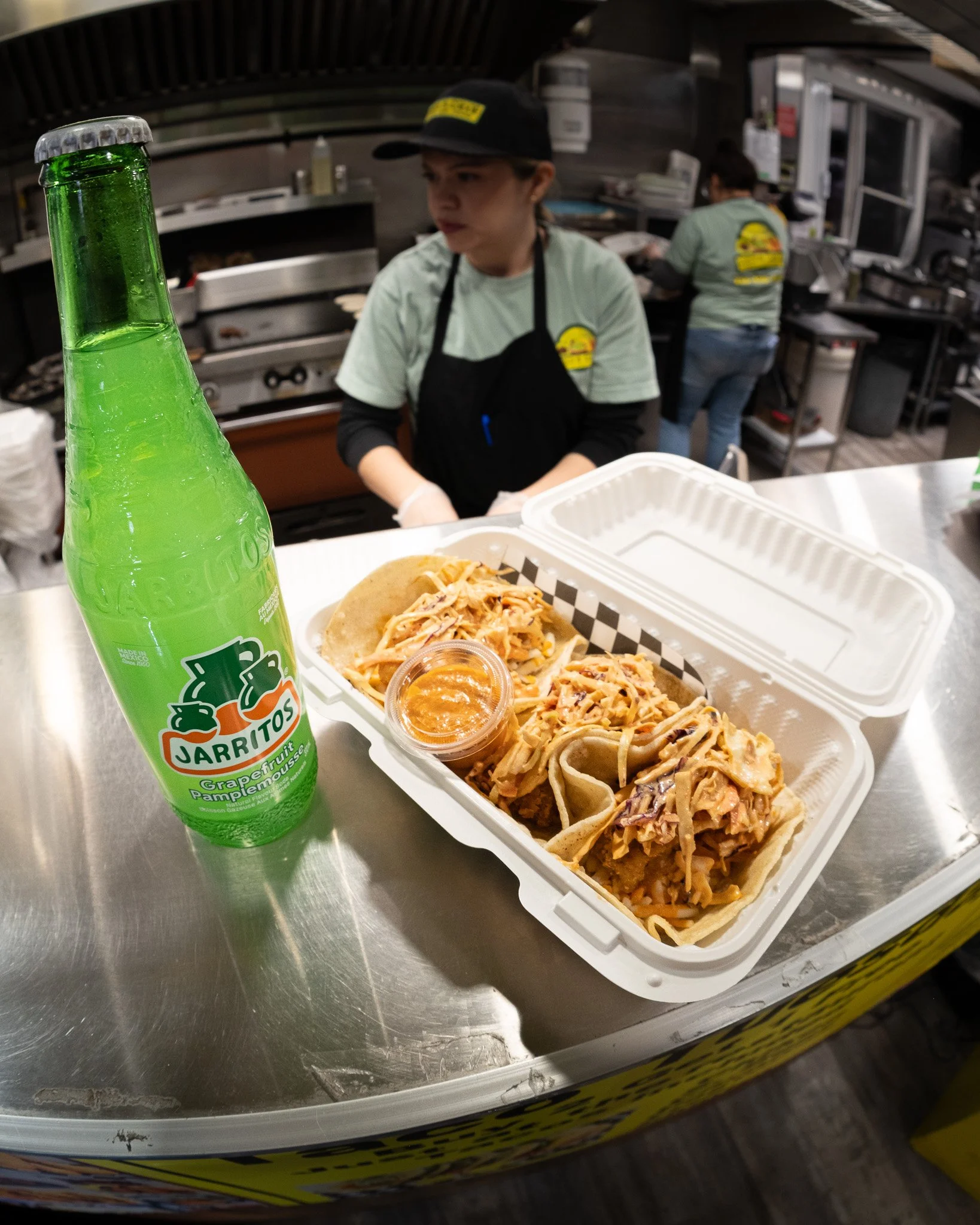 A green bottled Jarritos grapefruit soda and a takeout container with three tacos filled with shredded meat, topped with sauce, on a stainless steel counter at a restaurant.
