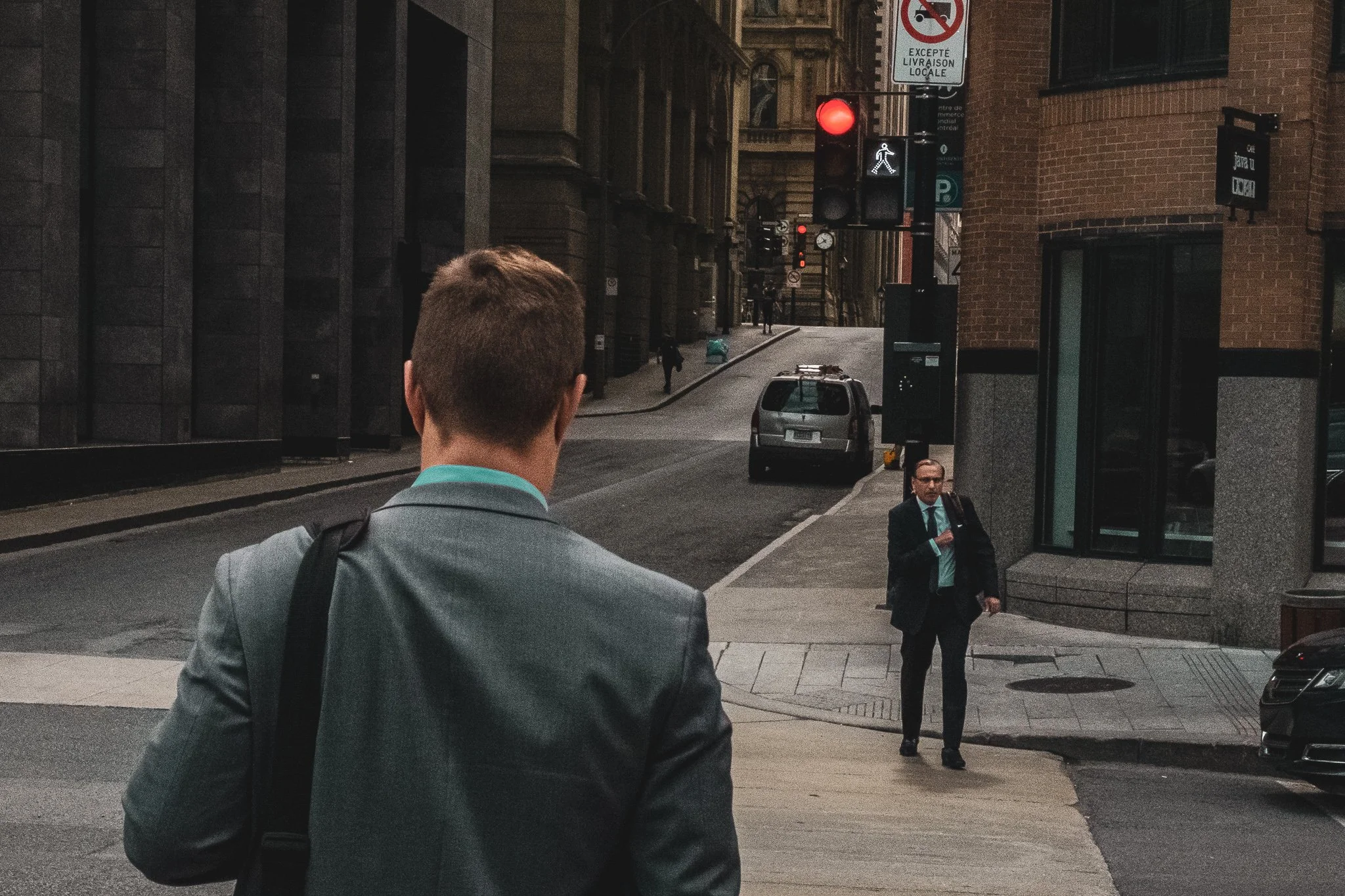 Two men in business attire in an urban city street, with one walking towards the camera and the other crossing the street, amidst tall buildings, parked cars, and traffic signals.