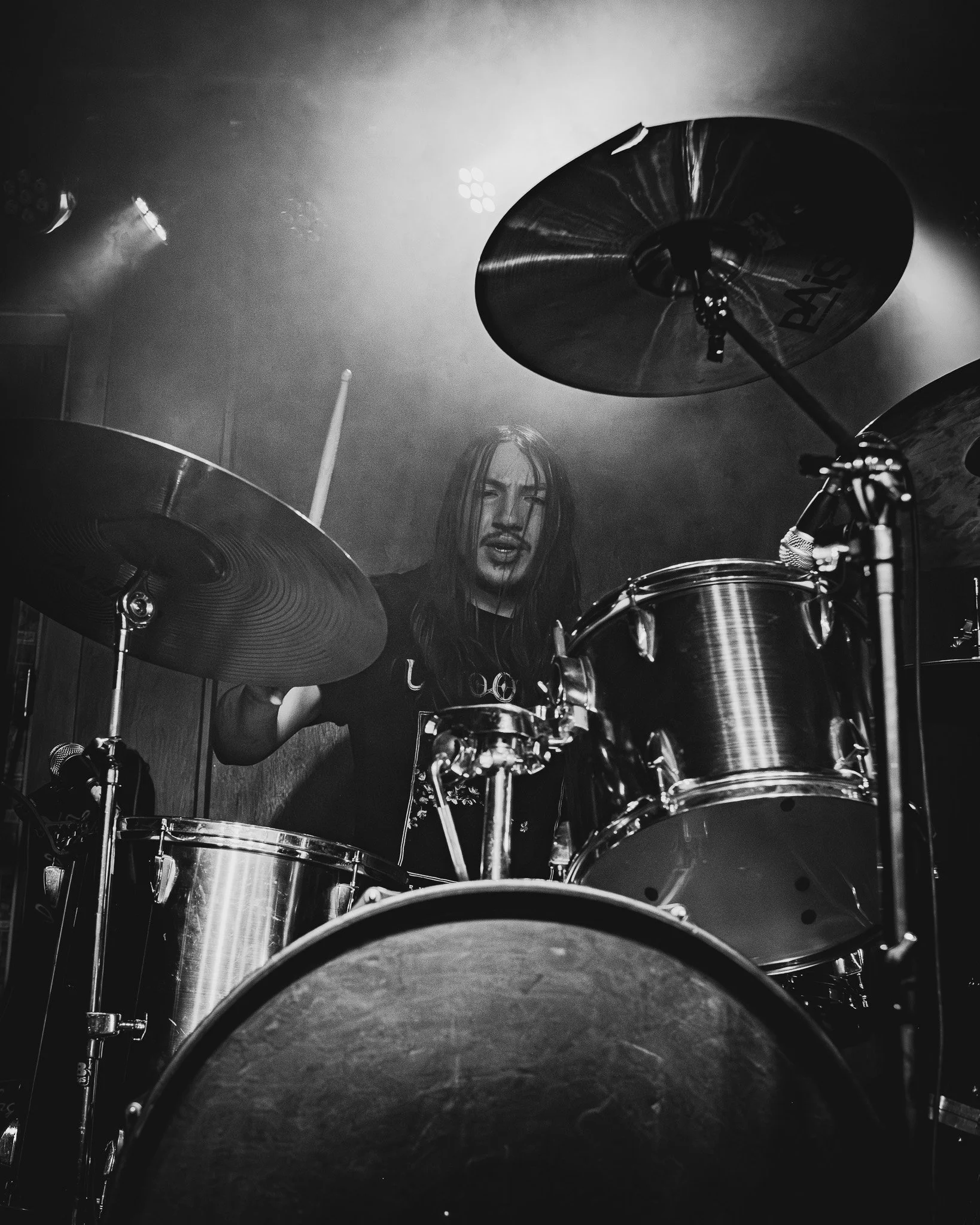 A black and white photo of a drummer with long hair playing a drum set on stage, with stage lights and smoke in the background.