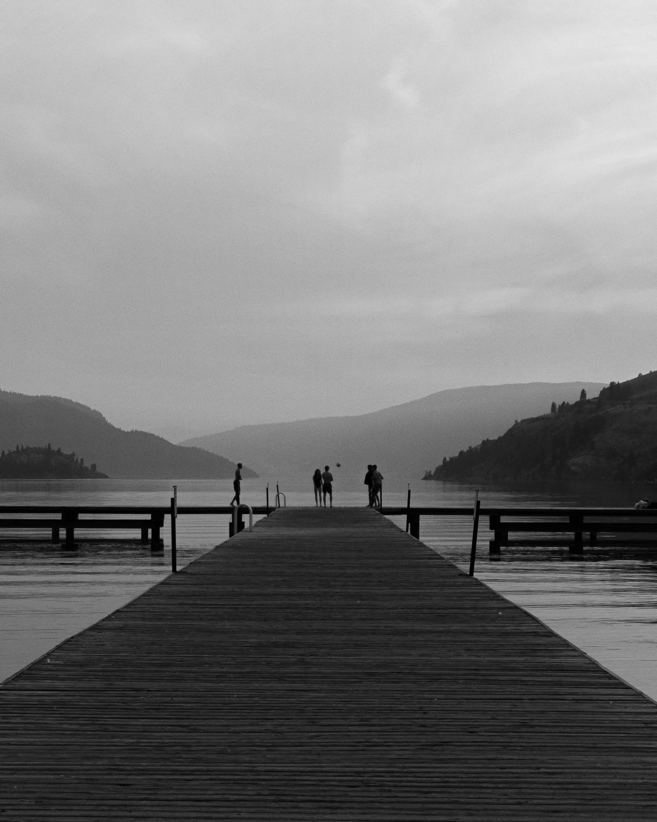 Black and white photo of a wooden dock extending into a calm lake, with four people standing at the end, surrounded by hills and mountains.