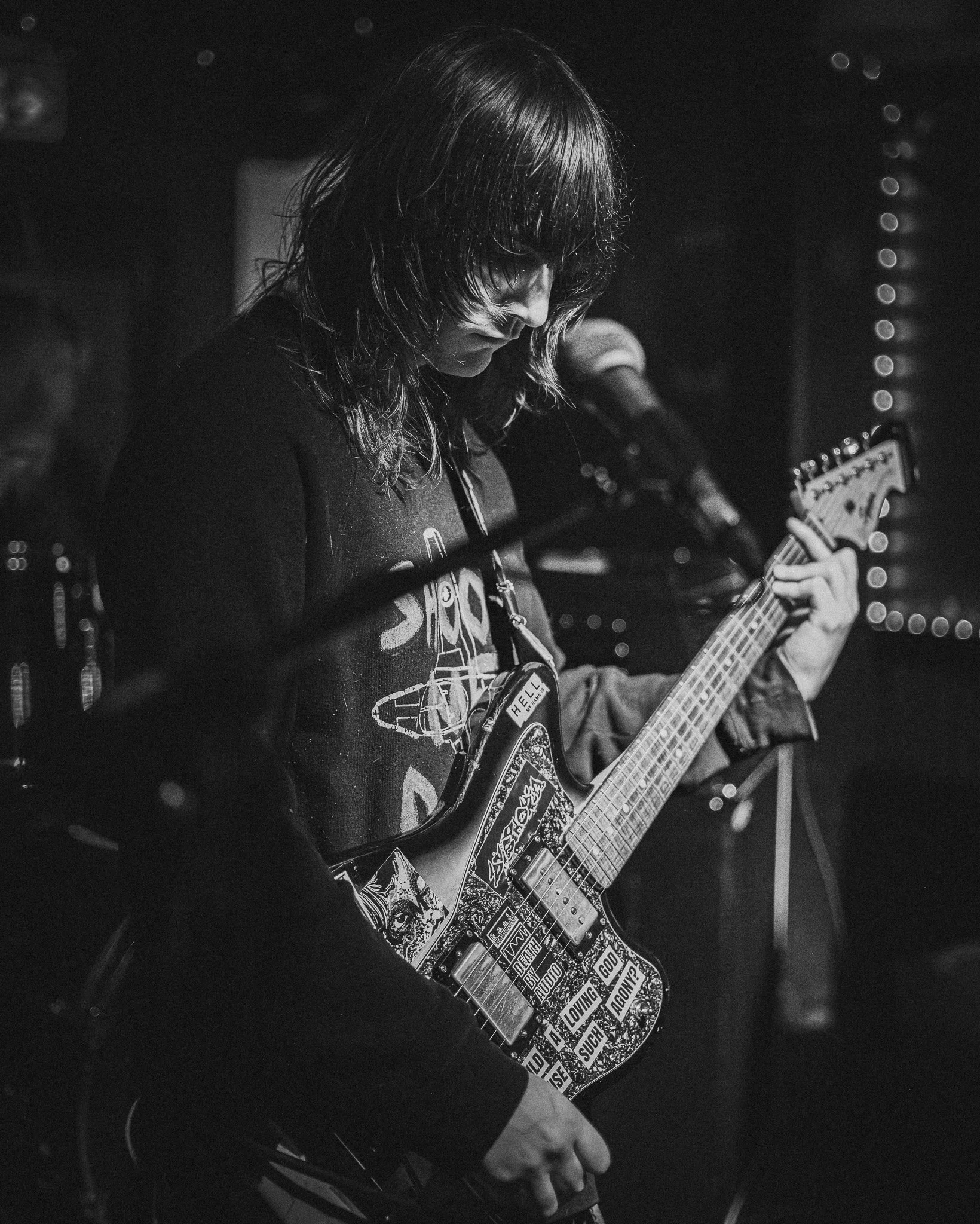 A person with shoulder-length hair playing an electric guitar on stage, with various stickers on the guitar.