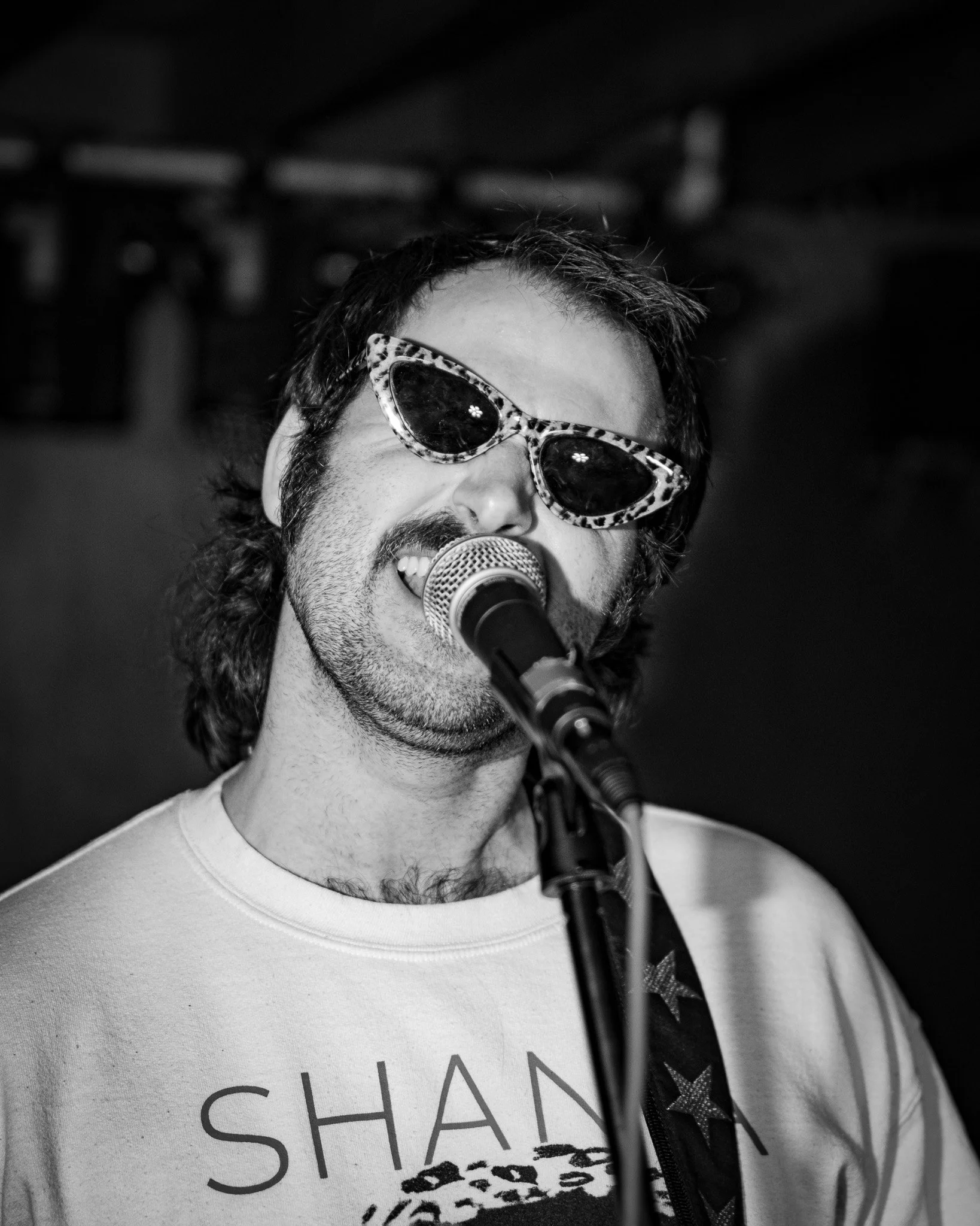 A man with dark hair, sunglasses with animal print frames, and a beard is singing into a microphone.