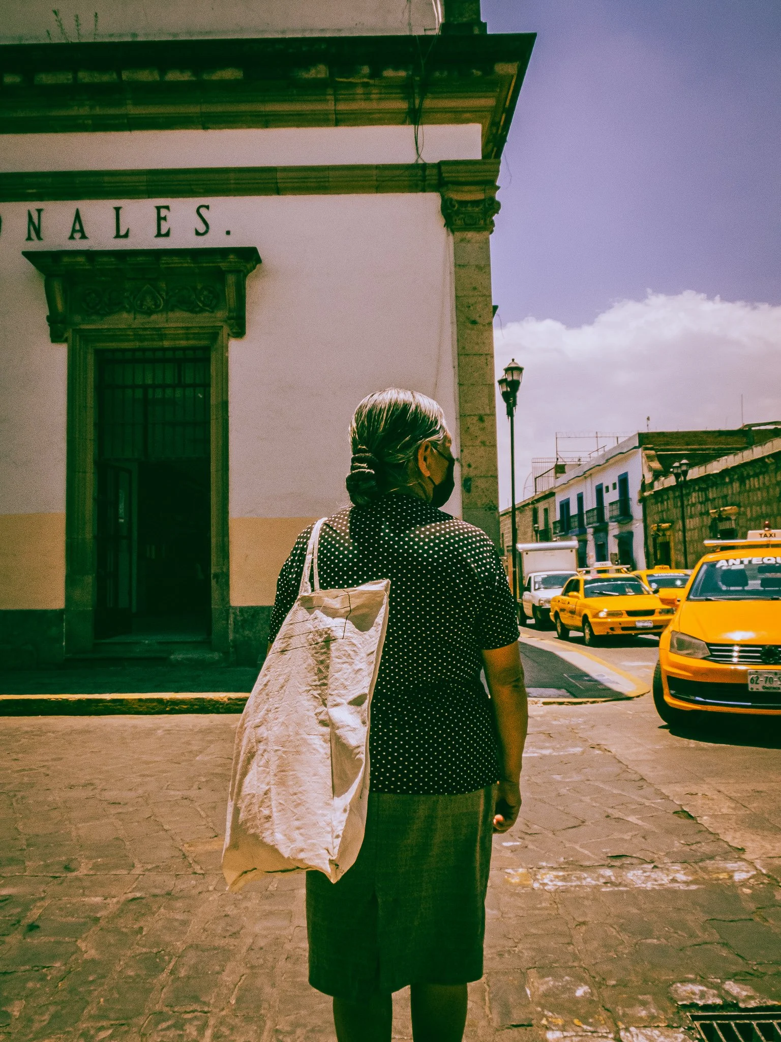 A woman with gray hair wearing a face mask, black polka-dot top, and long black skirt, stands on a cobblestone street with yellow taxis and historic buildings in the background.
