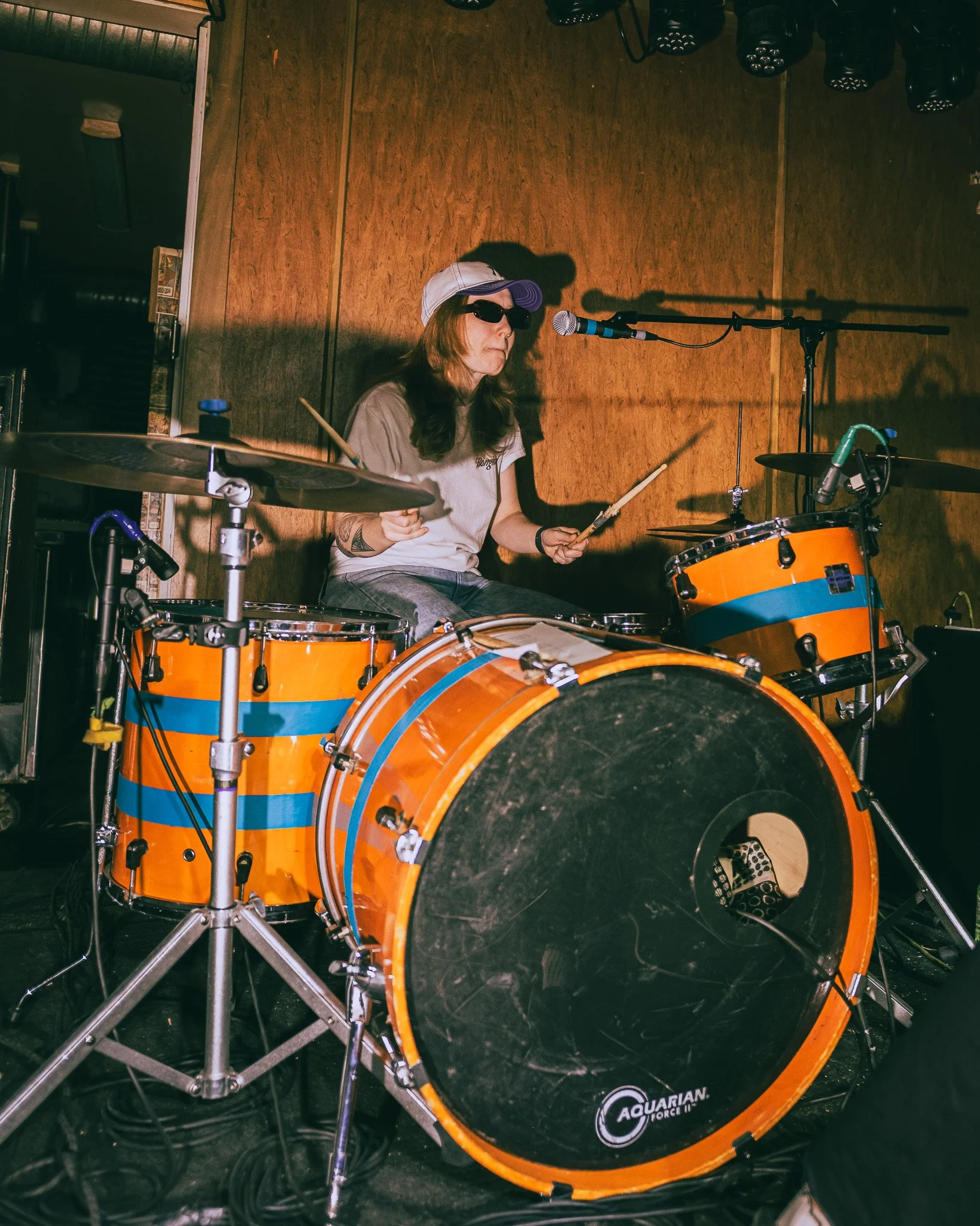 A woman wearing sunglasses and a hat playing a bright orange drum set with blue stripes in a performance space with wooden walls.