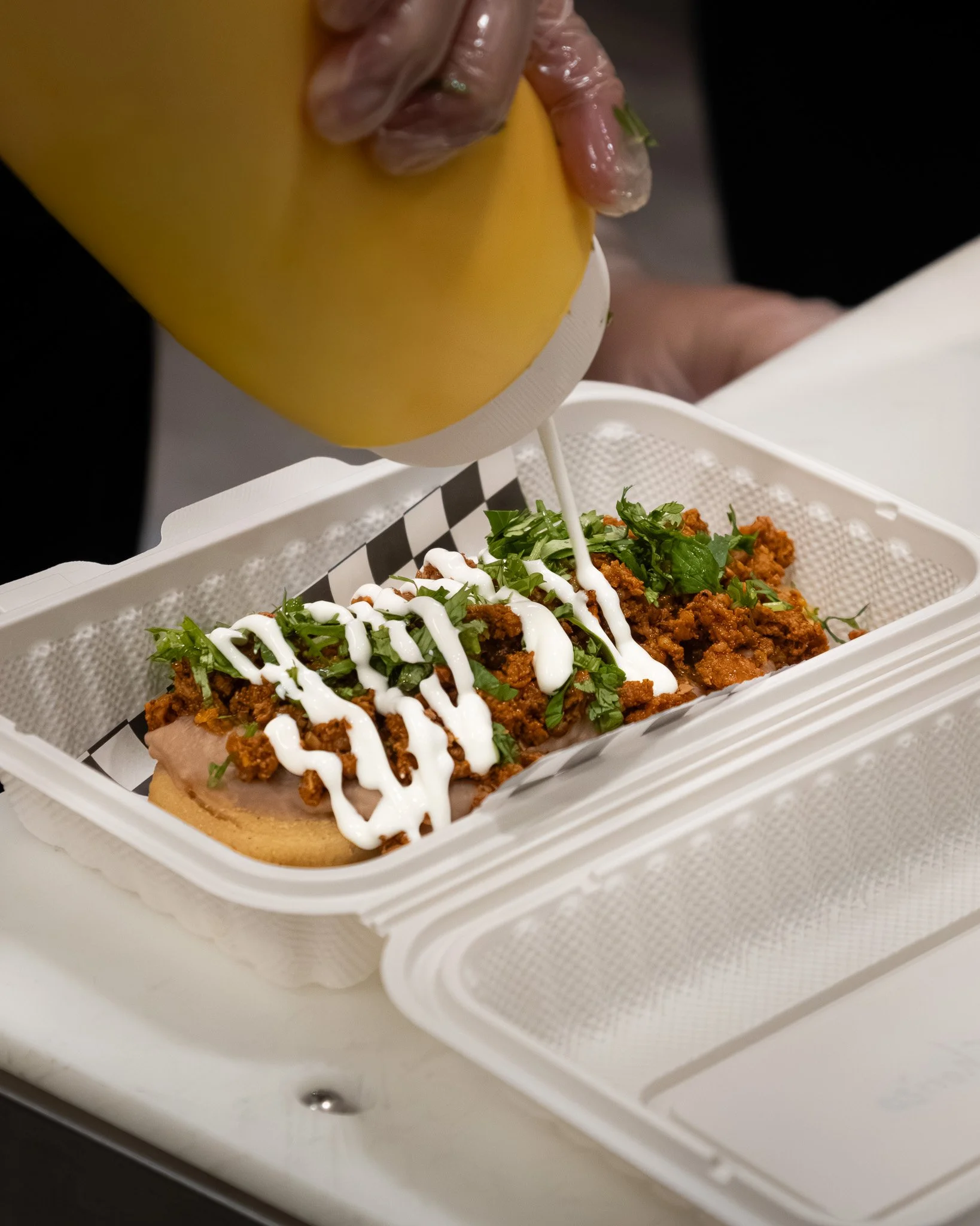 Hot dog in a paper tray being topped with mustard, mayonnaise, chopped cilantro, and crushed chips.