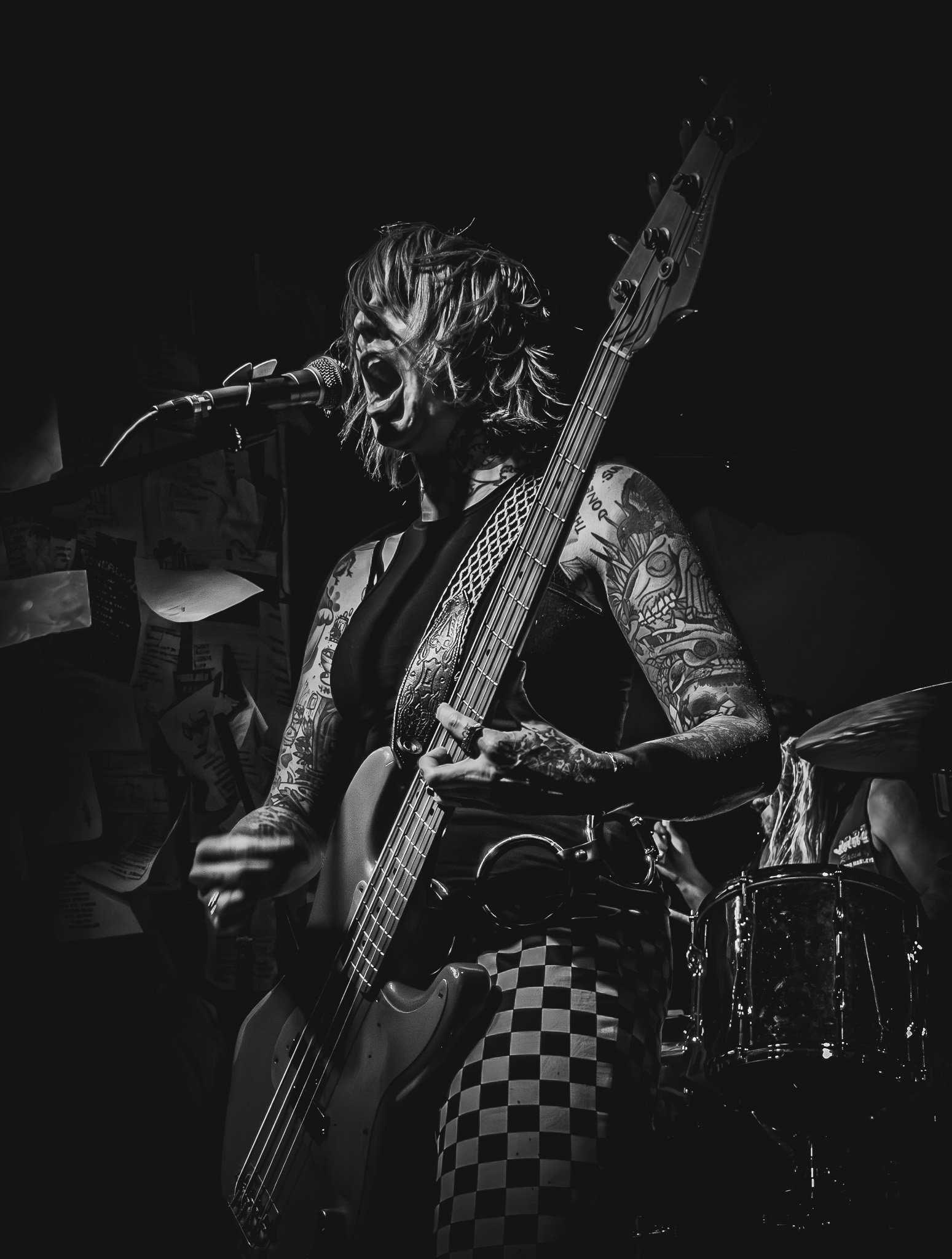Black and white photo of a female musician performing on stage, singing into a microphone and playing an electric bass guitar, with tattoos on her arms and checkered pants, in a dimly lit setting.