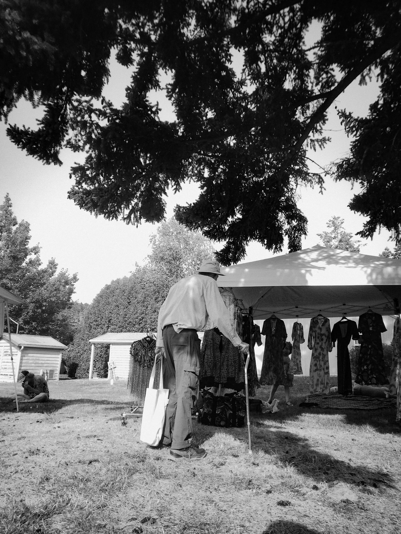 A man wearing a hat and sunglasses with a cane shopping at an outdoor market under a tent with clothing on display, with trees and small buildings in the background.
