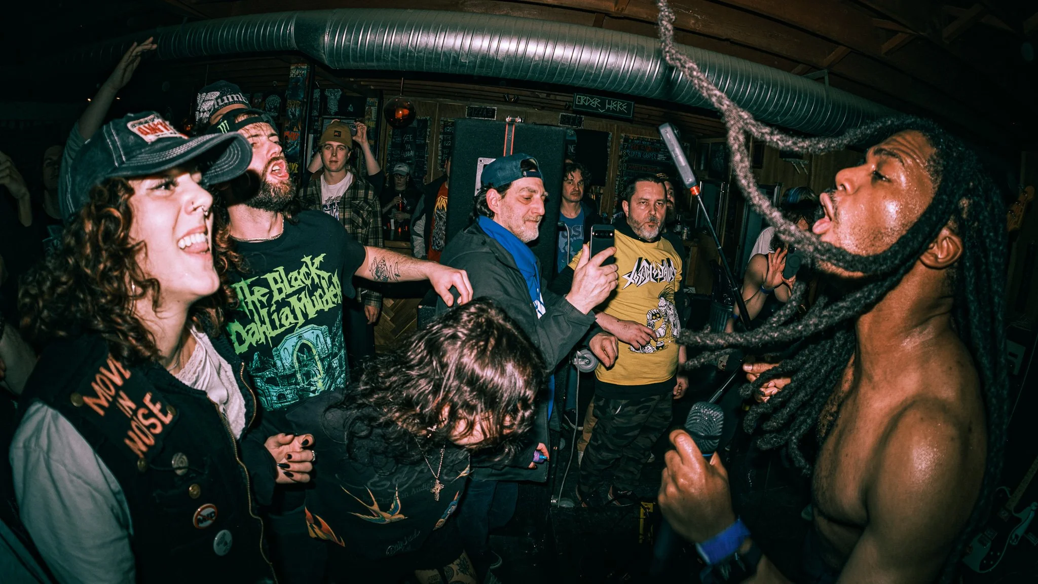 A live music performance at a small indoor venue with a shirtless lead singer with dreadlocks singing into a microphone, surrounded by a crowd of enthusiastic fans, some with phones recording.